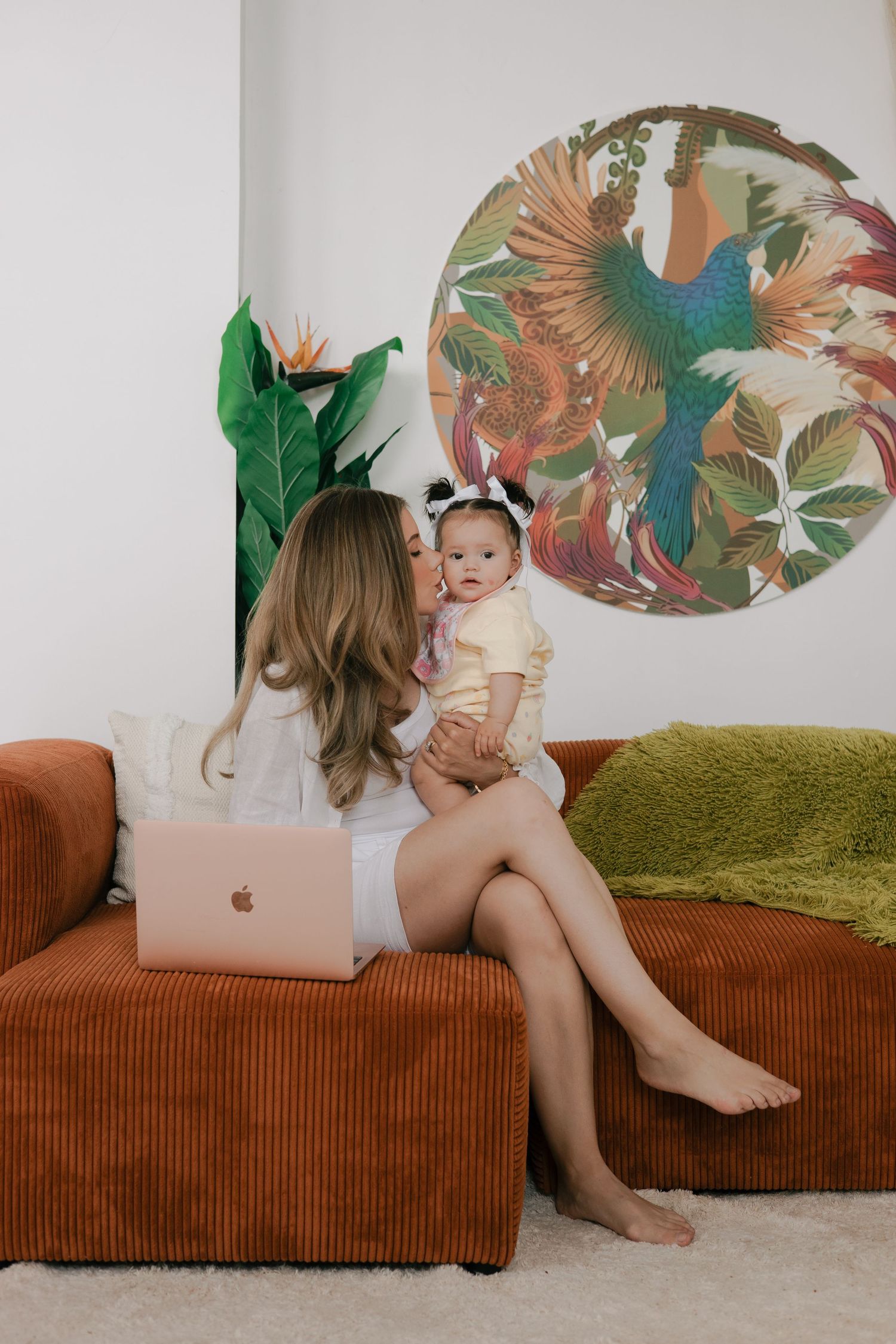 A person sits on a rust-colored corduroy couch with a laptop beneath tropical bird wall art in a modern living space.