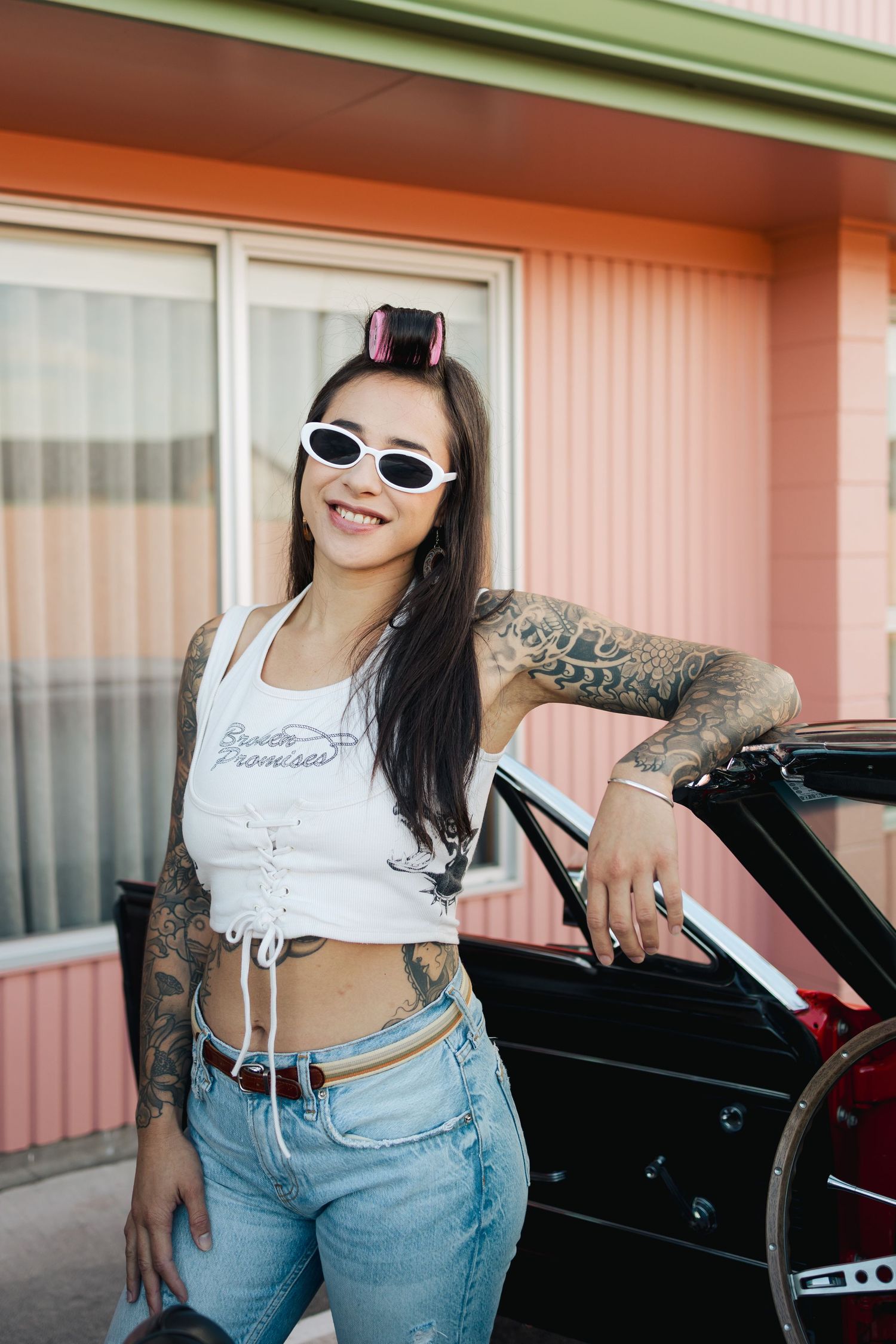 Woman in sunglasses and crop top posing near vintage car at retro motel.