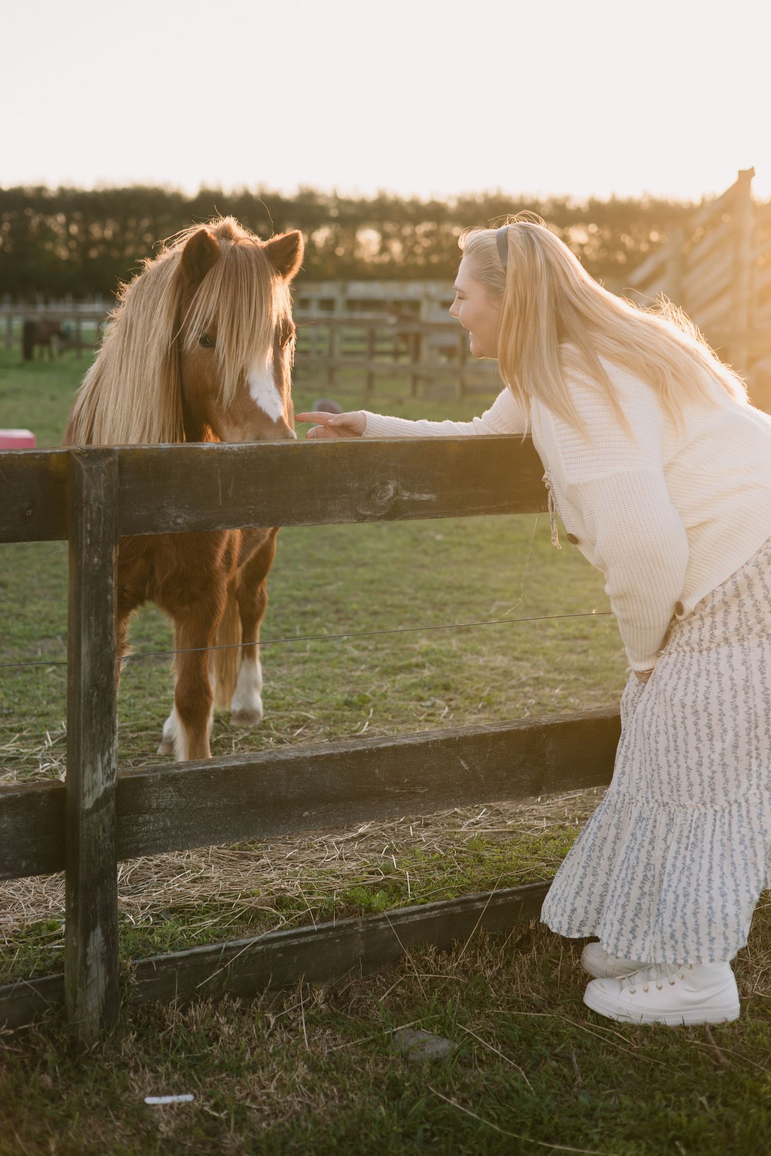 Woman in white sweater leaning on wooden fence to pet a light brown horse at sunset.