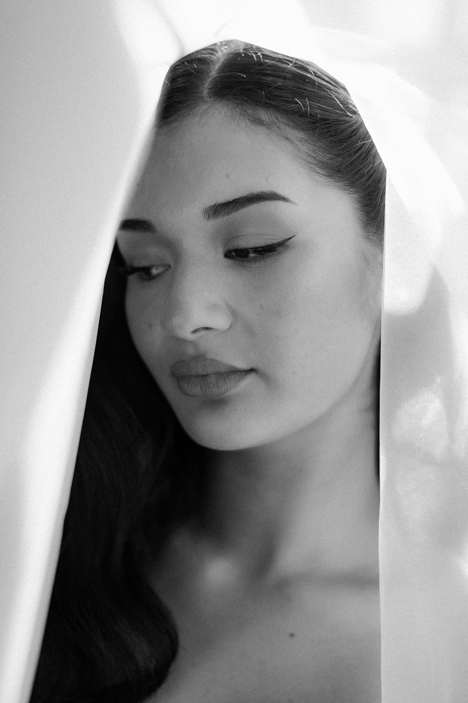 Black and white portrait of a young woman with winged eyeliner peeking through white fabric, looking down.