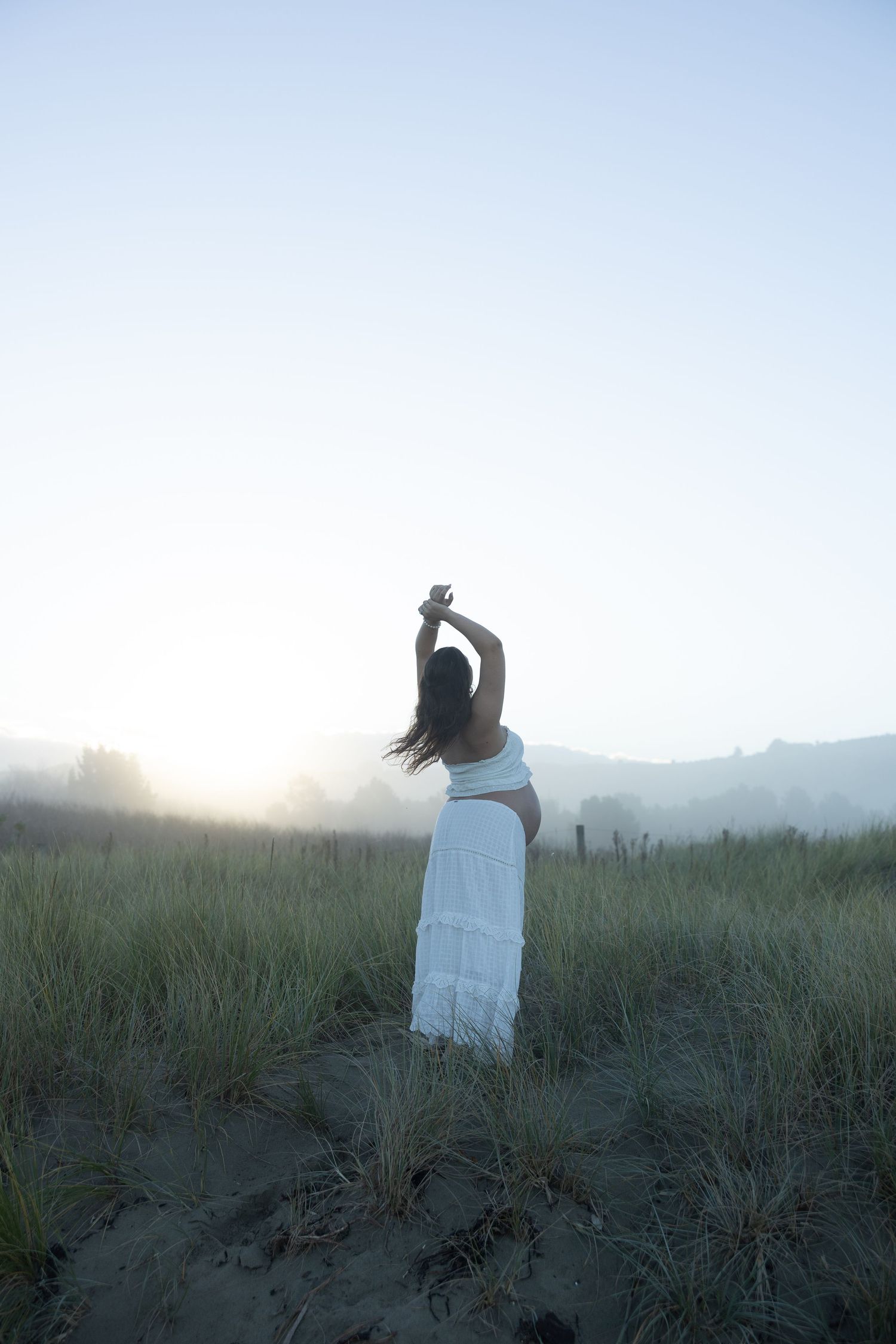 Woman in white dress standing in misty field at sunrise with arms raised gracefully above her head.