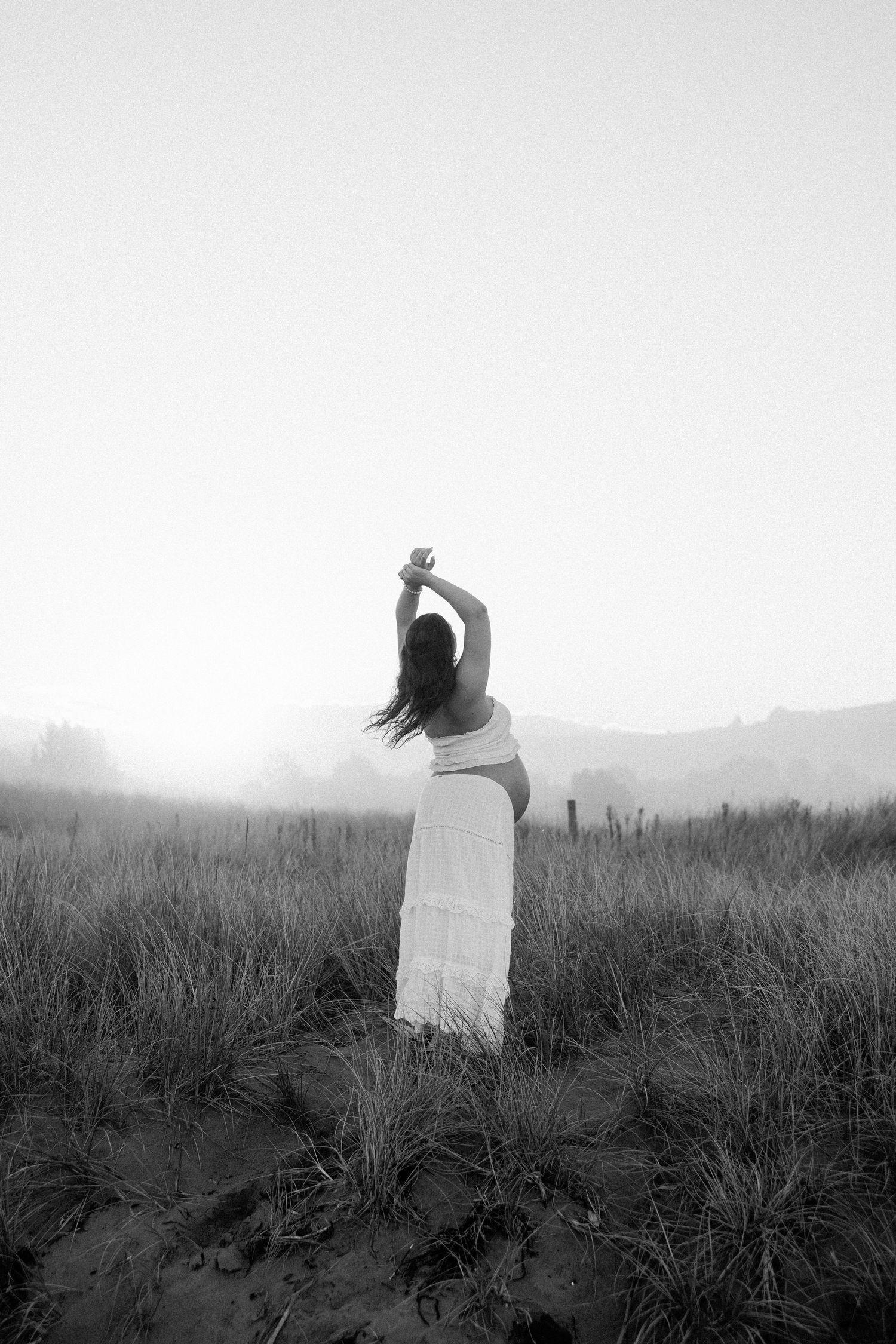 Black and white photo of a woman in a white dress standing in a misty field with her arms raised gracefully.