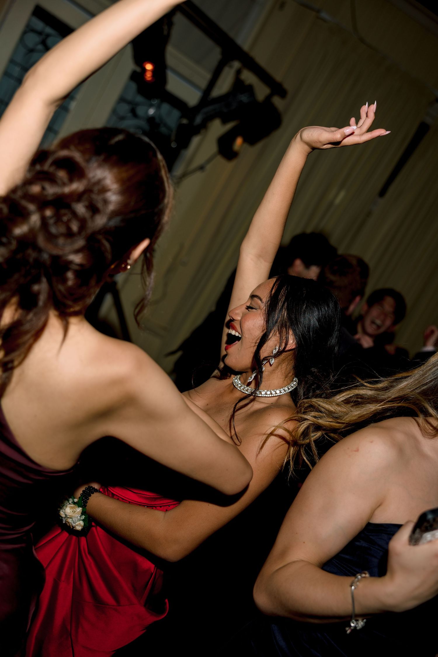 Young women in formal dresses dancing joyfully at a party, one laughing with arms raised.