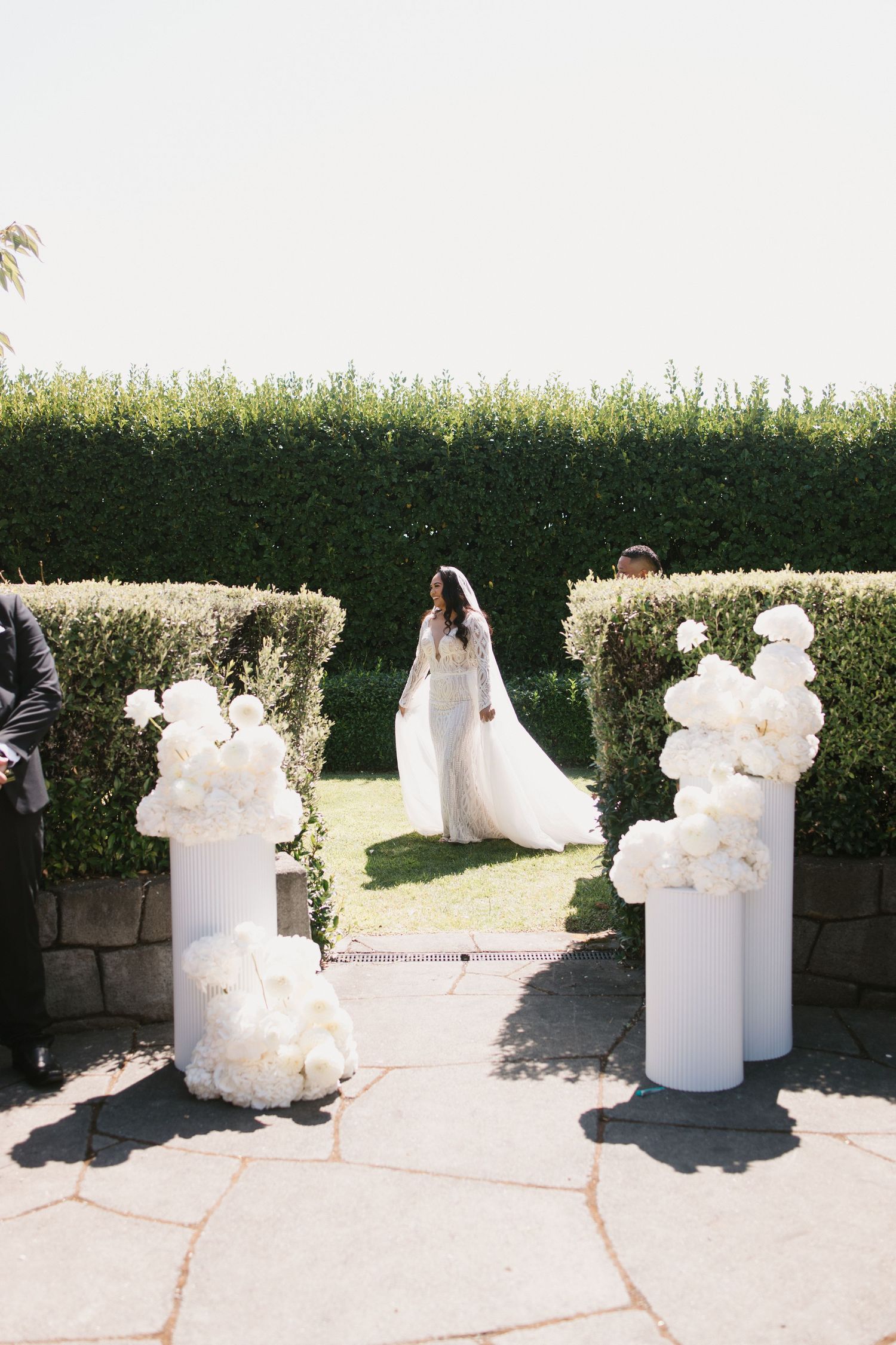 A sequence of white plaster or stone sculptures displayed in a garden setting against a green hedge backdrop.