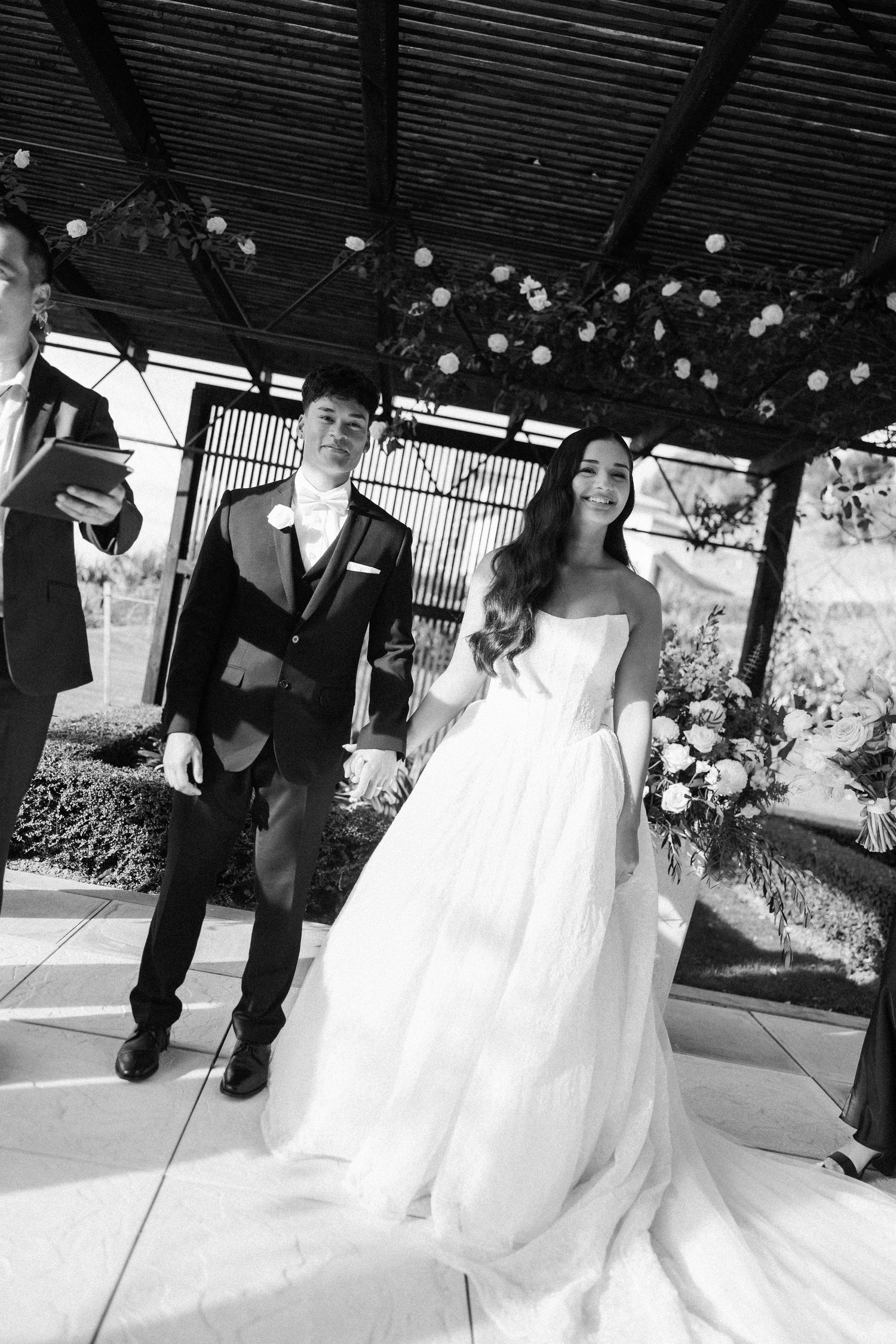 Black and white photo of a smiling bride in a strapless gown holding bouquet beside groom in suit at outdoor wedding ceremony.
