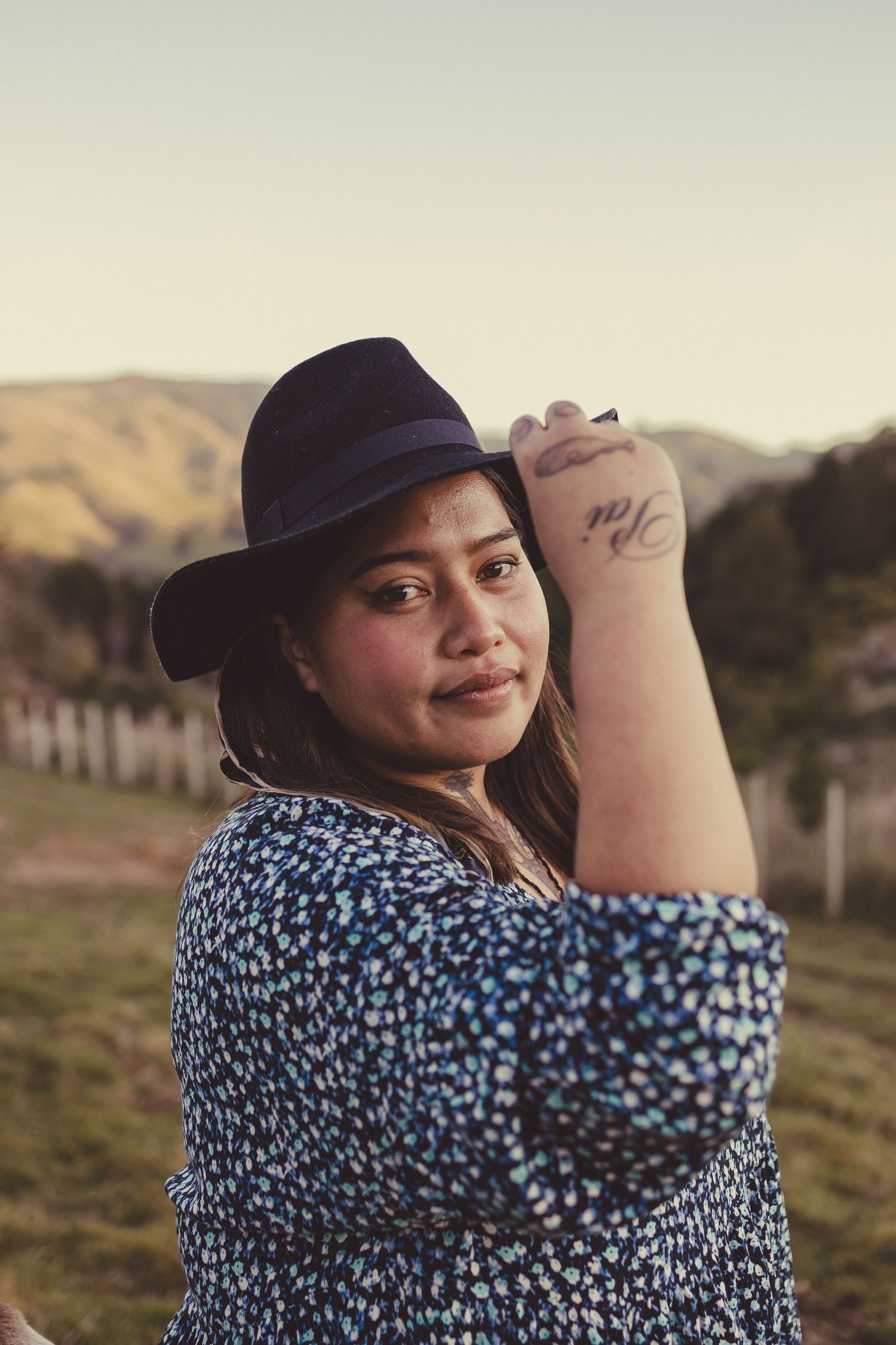 Woman in blue floral dress and black fedora hat posing outdoors with hills in background, showing wrist tattoo.