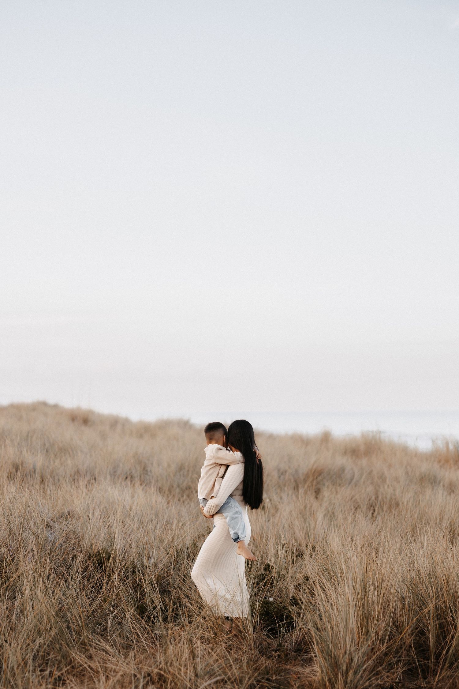 A woman in a cream dress holds a baby in a golden grass field by the ocean at dusk.