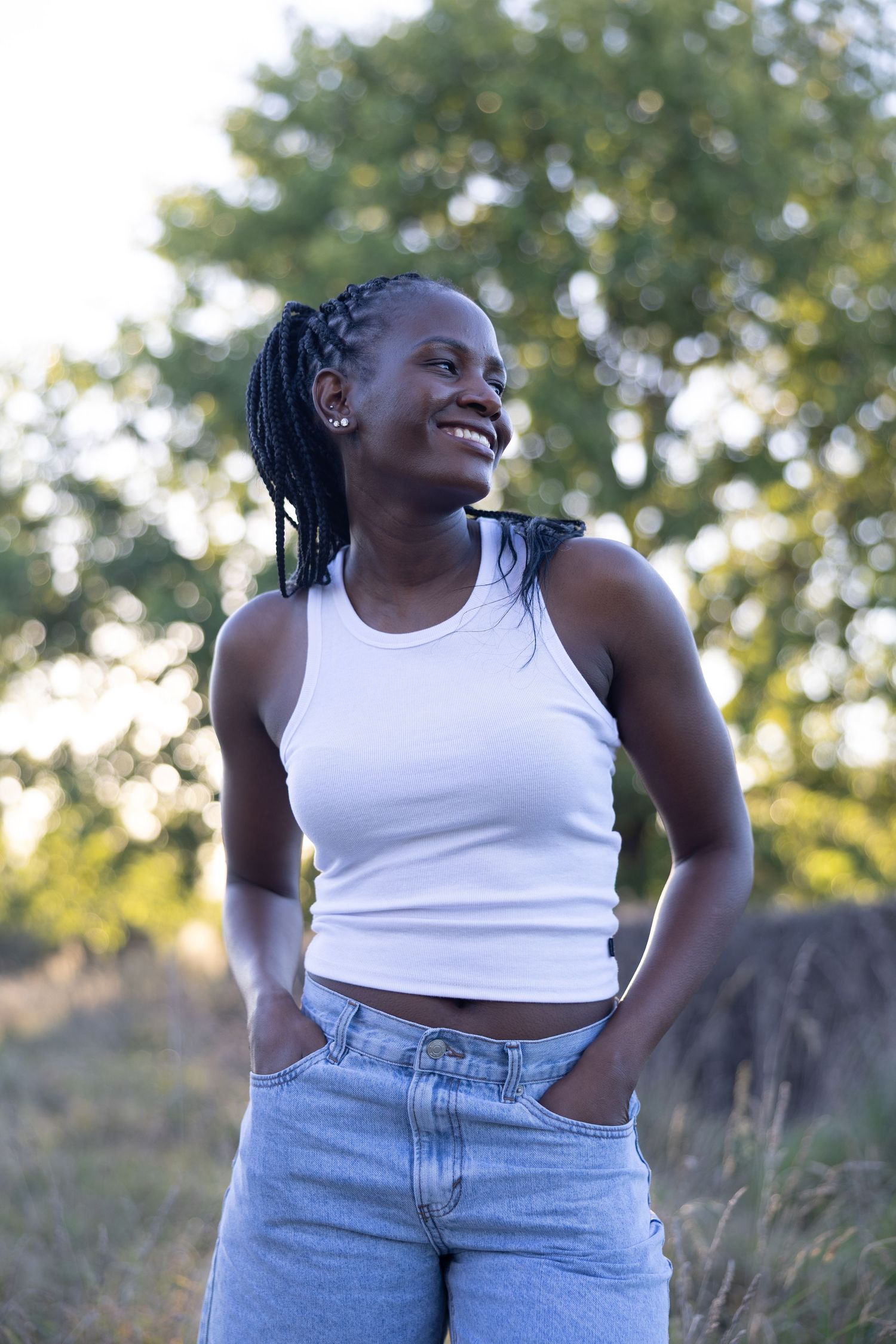 Smiling young Black woman with braids wearing white crop top and jeans standing outdoors in natural setting.