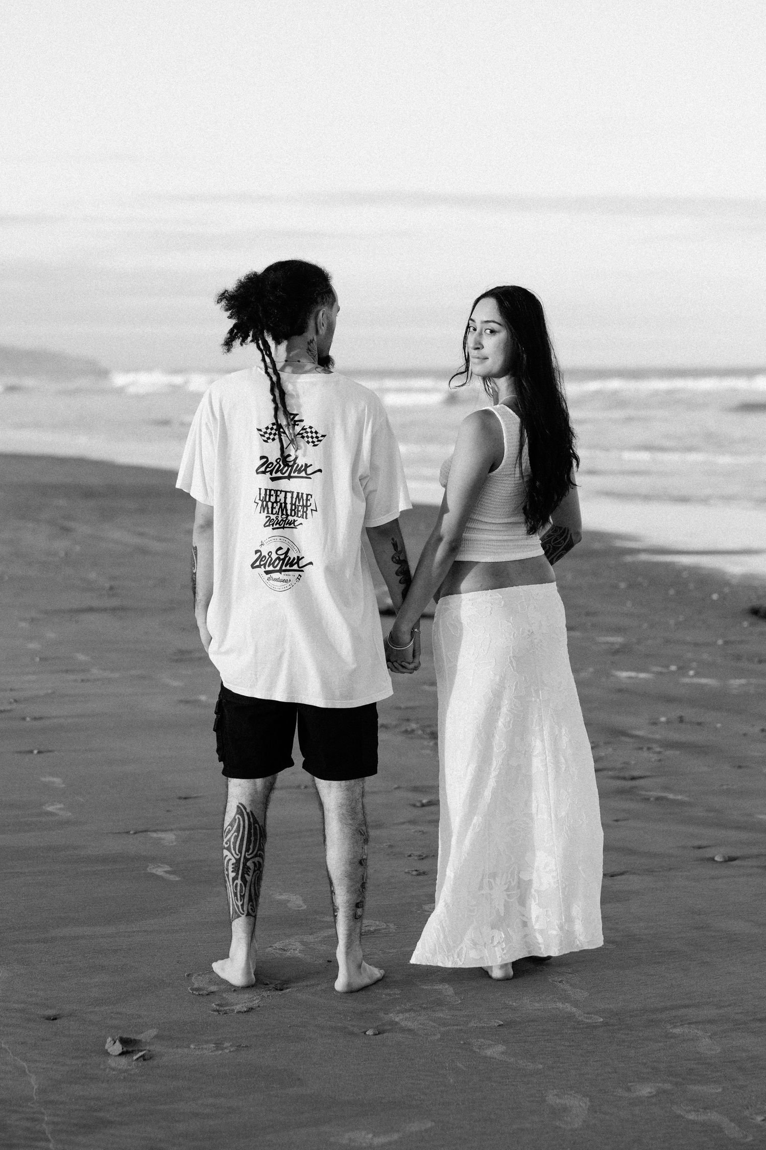 Black and white photo of a couple holding hands on a beach, man with tattoos and braids, woman in white skirt.