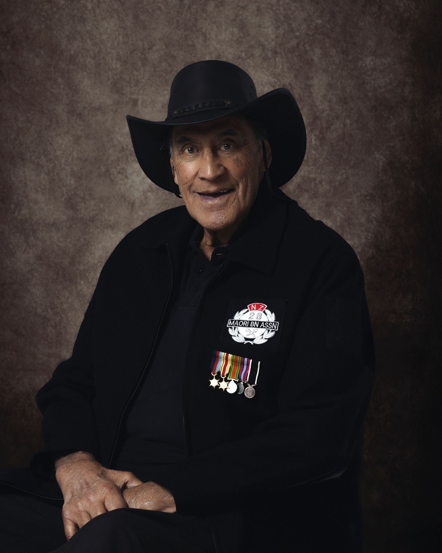 Elderly veteran in black cowboy hat wearing military medals and a badge, posed for a formal portrait photo.