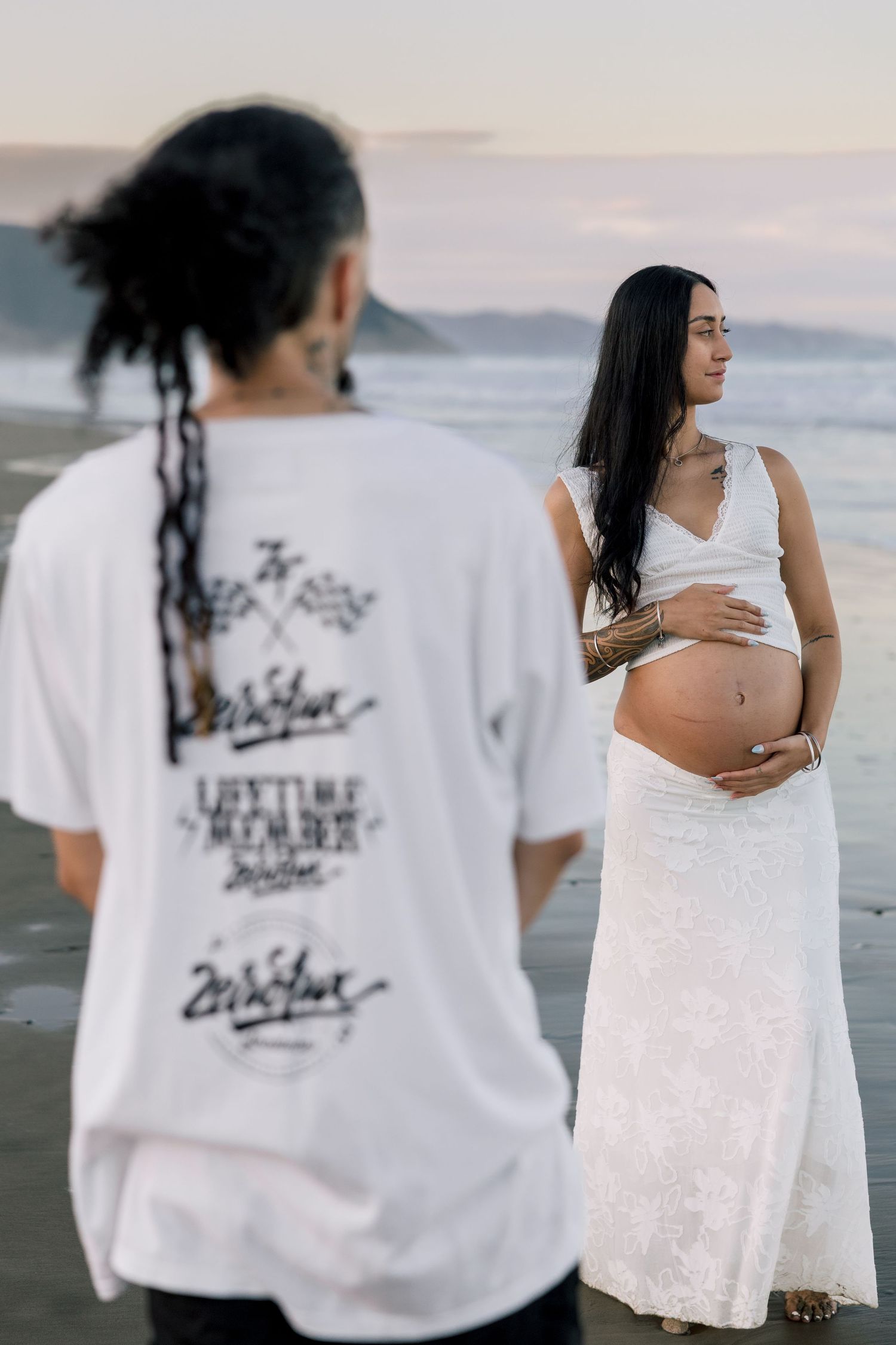 Pregnant woman in white lace dress on beach, partner with dreadlocks and graphic tee in foreground.
