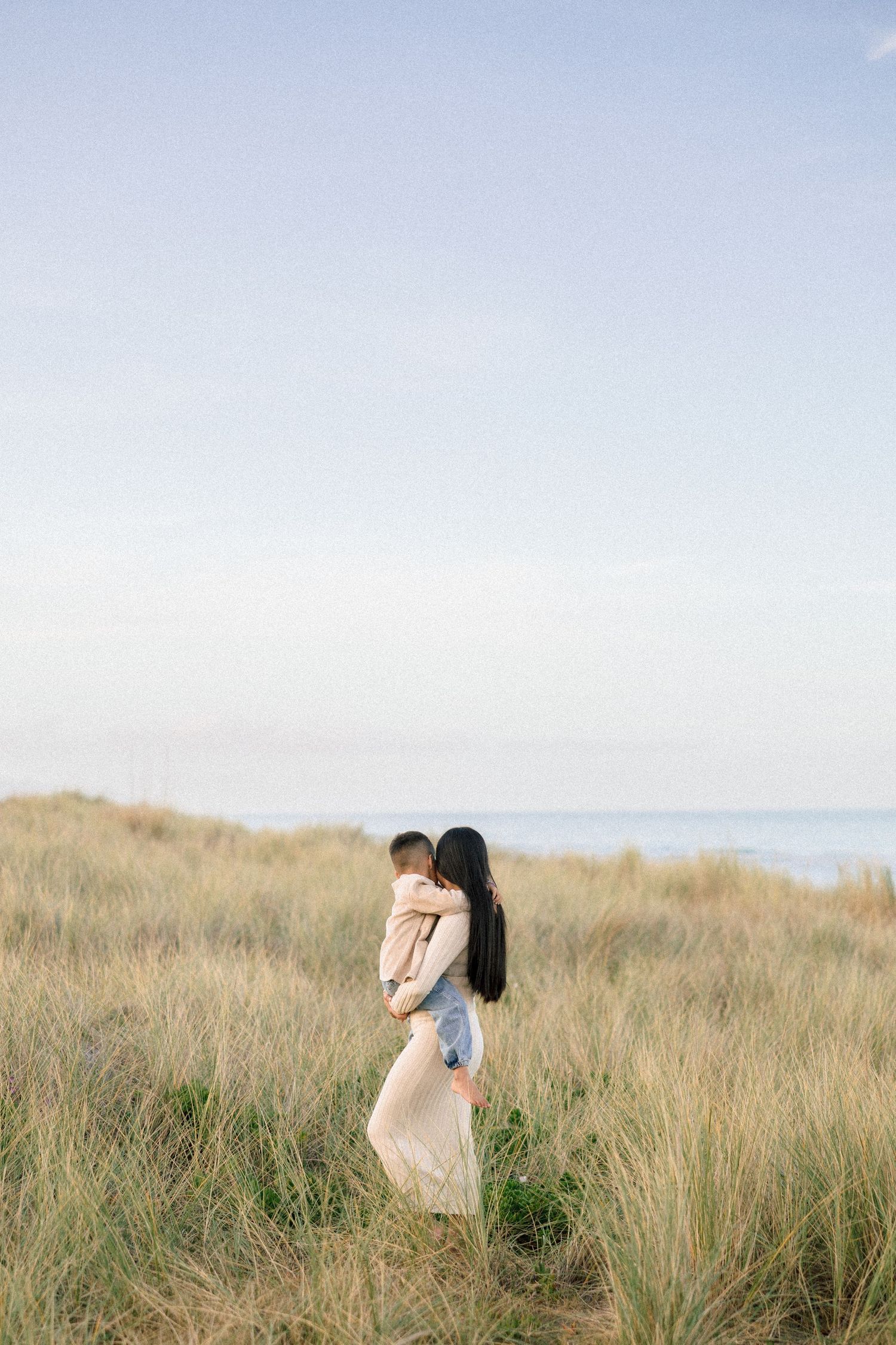 Mother with long dark hair holds young child in golden beach grass dunes under soft sky.