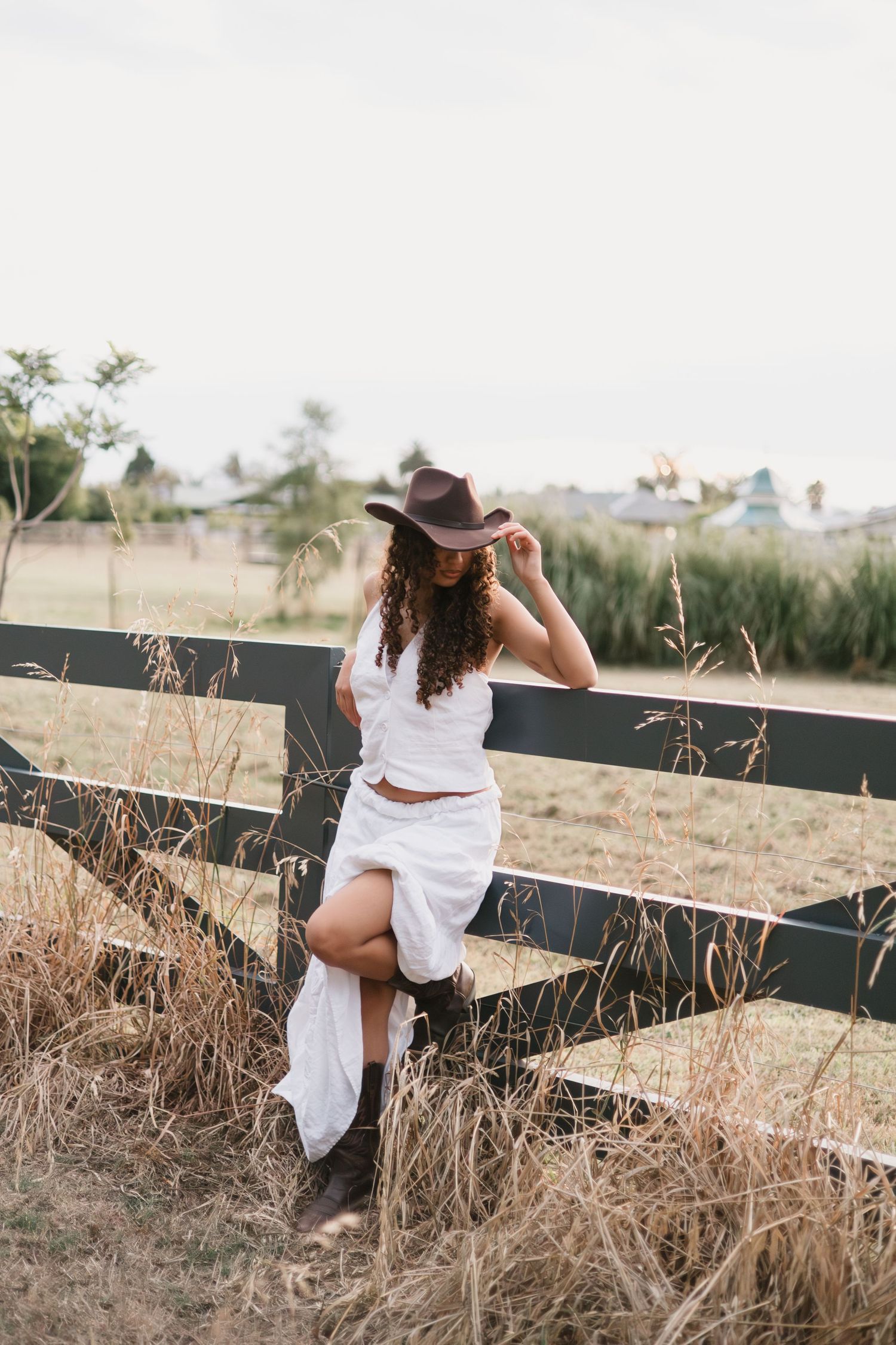 A cowgirl sits on a wooden fence at sunset in a white dress and boots.