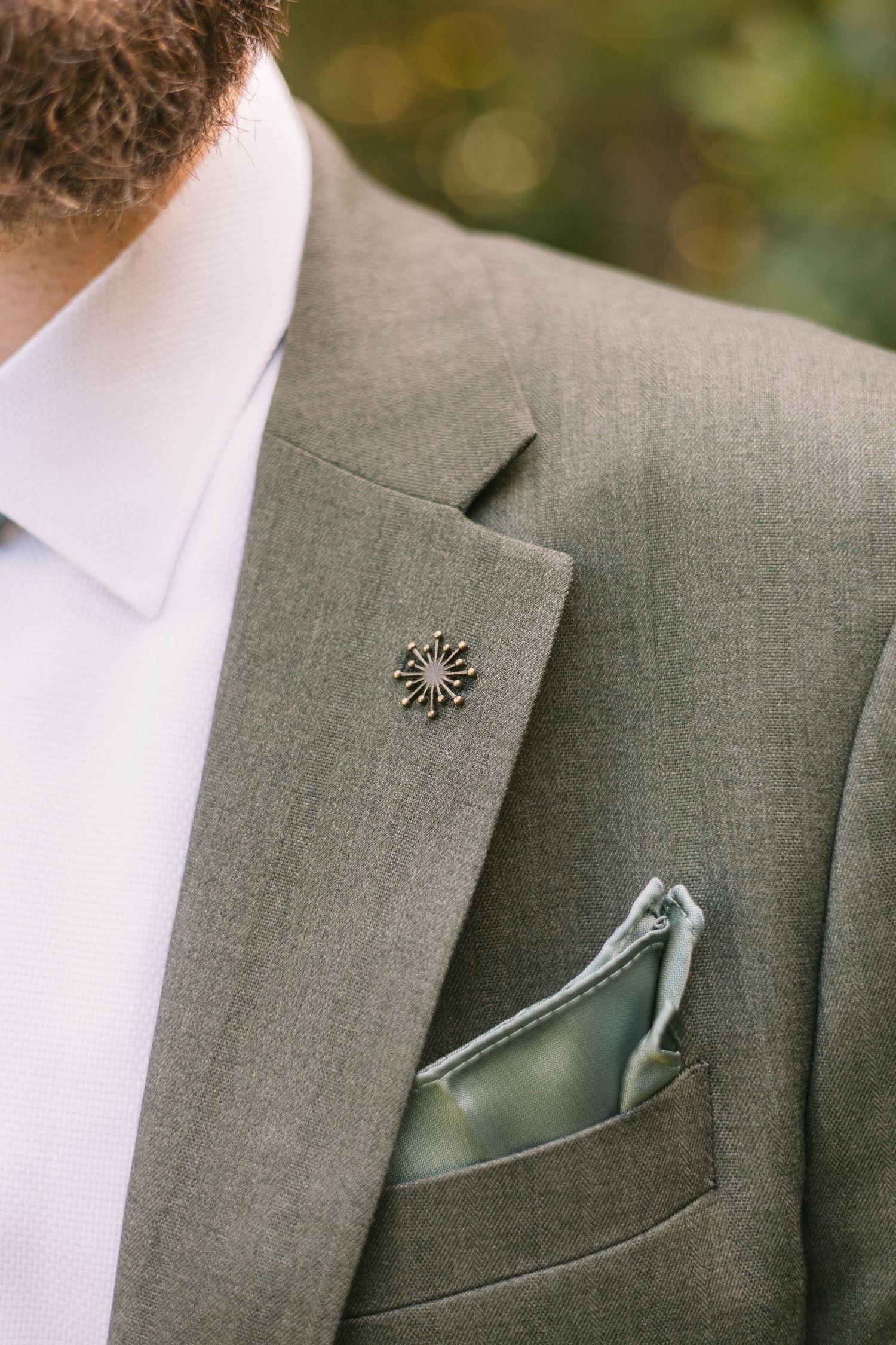 Close-up view of a grey suit lapel with a decorative pin and sage green pocket square.