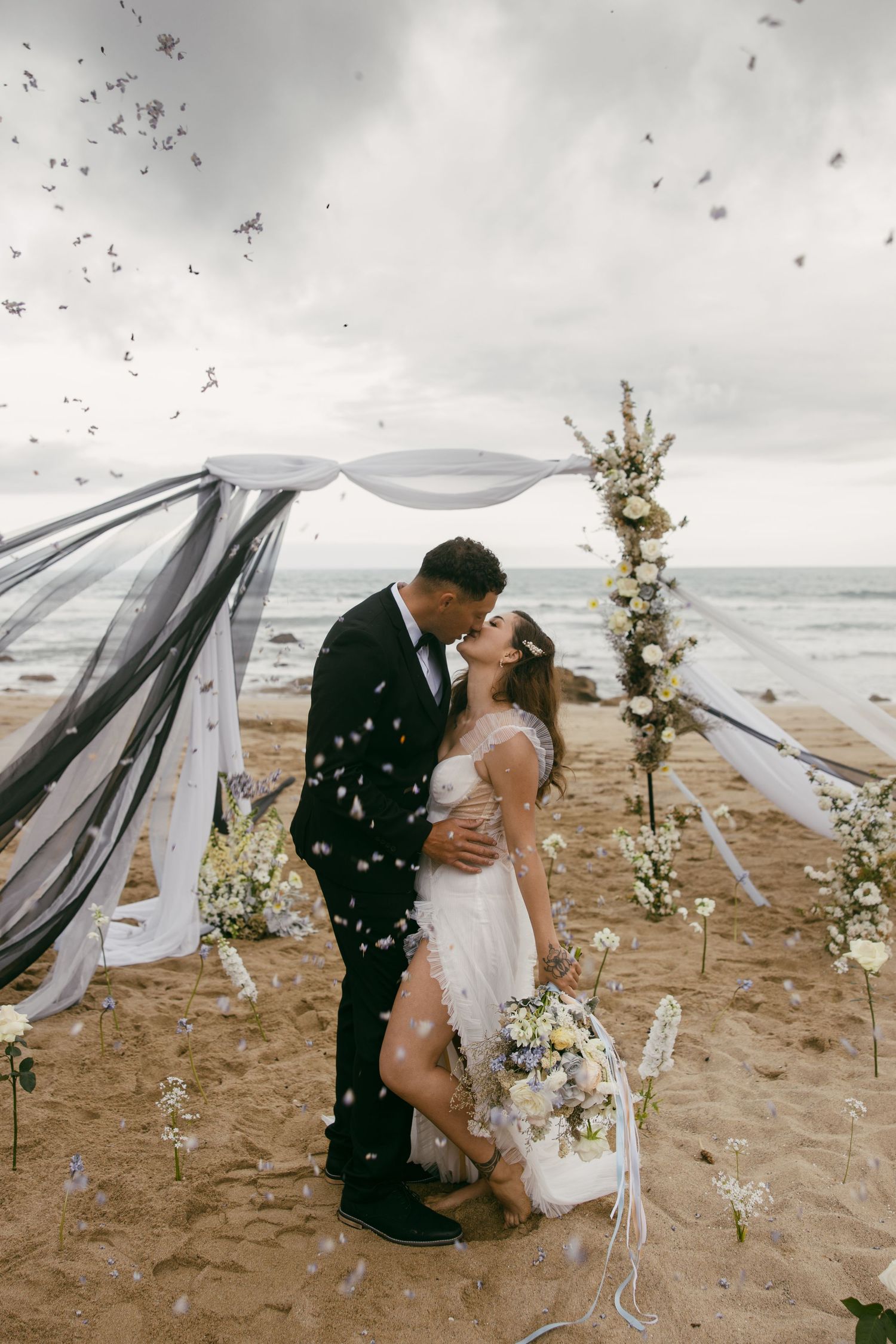 Romantic couple shares tender moment on beach with flowing white fabric and floral decorations during cloudy day ceremony.
