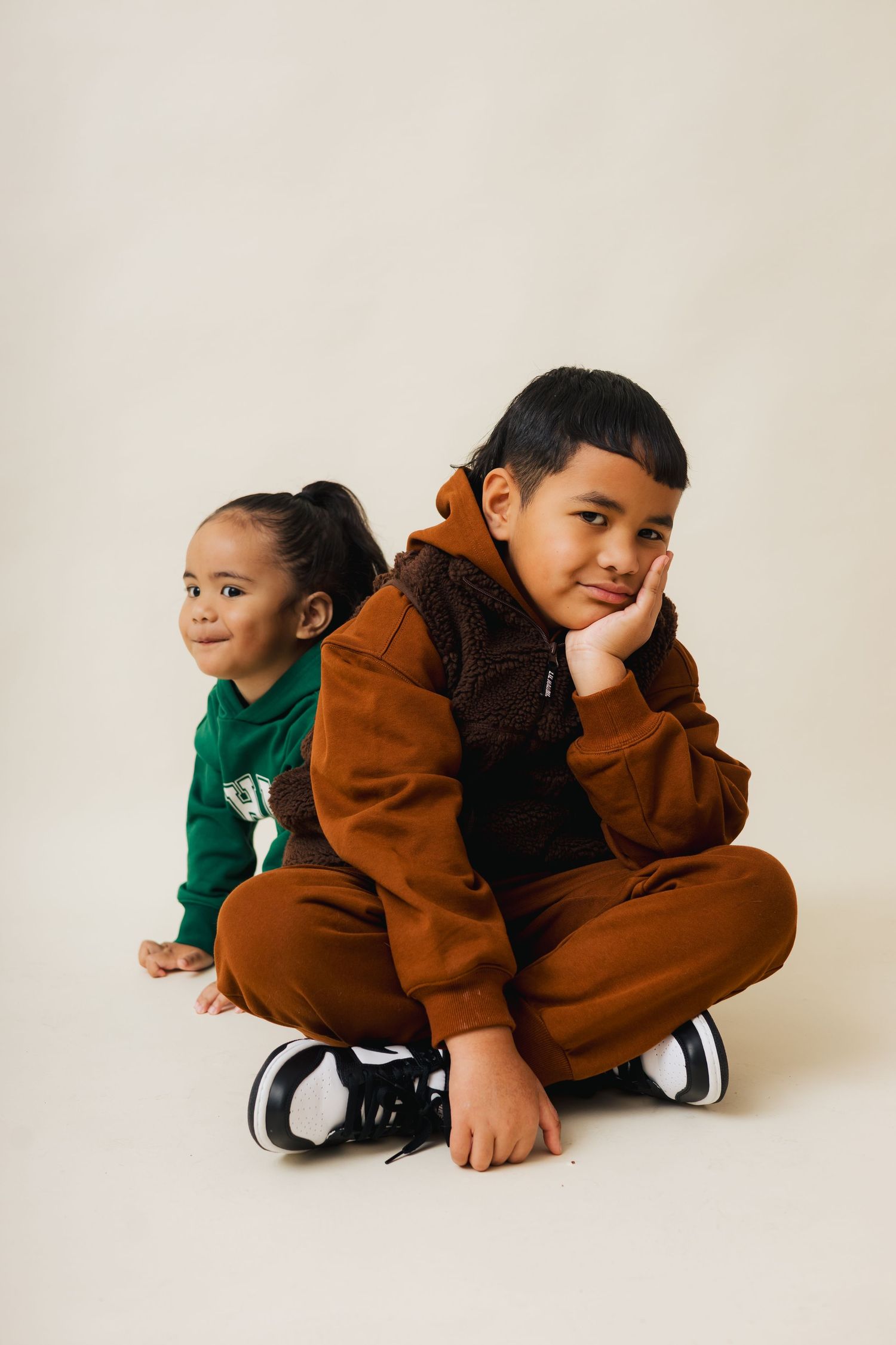 Two children sit on a white floor wearing casual clothing in brown and green tones.