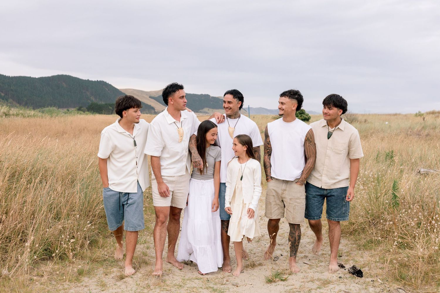 A group in coordinated white and beige outfits walks together through a scenic grassy field at sunset.