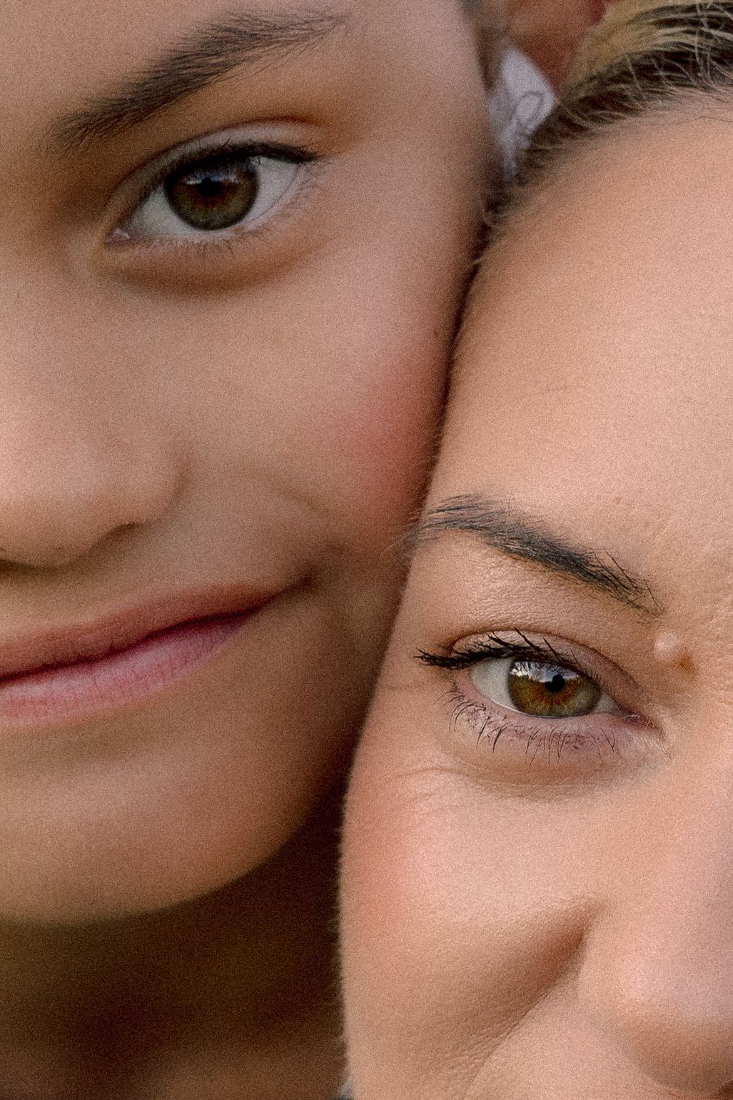 Close-up portrait of two young women with hazel eyes and glowing skin posing cheek to cheek.