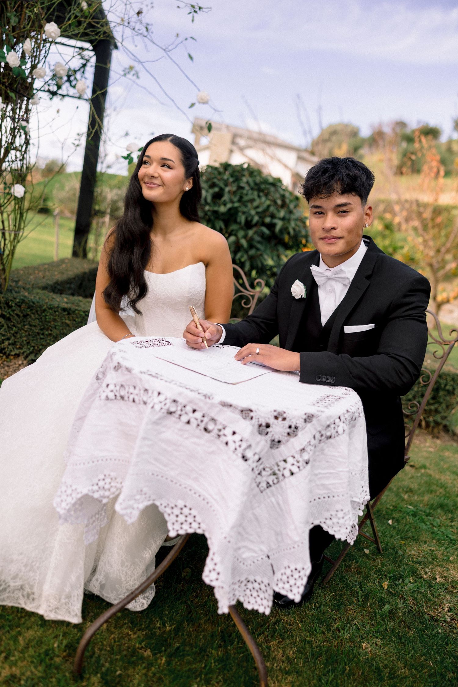 Bride and groom signing wedding registry at lace-covered table in outdoor garden ceremony.
