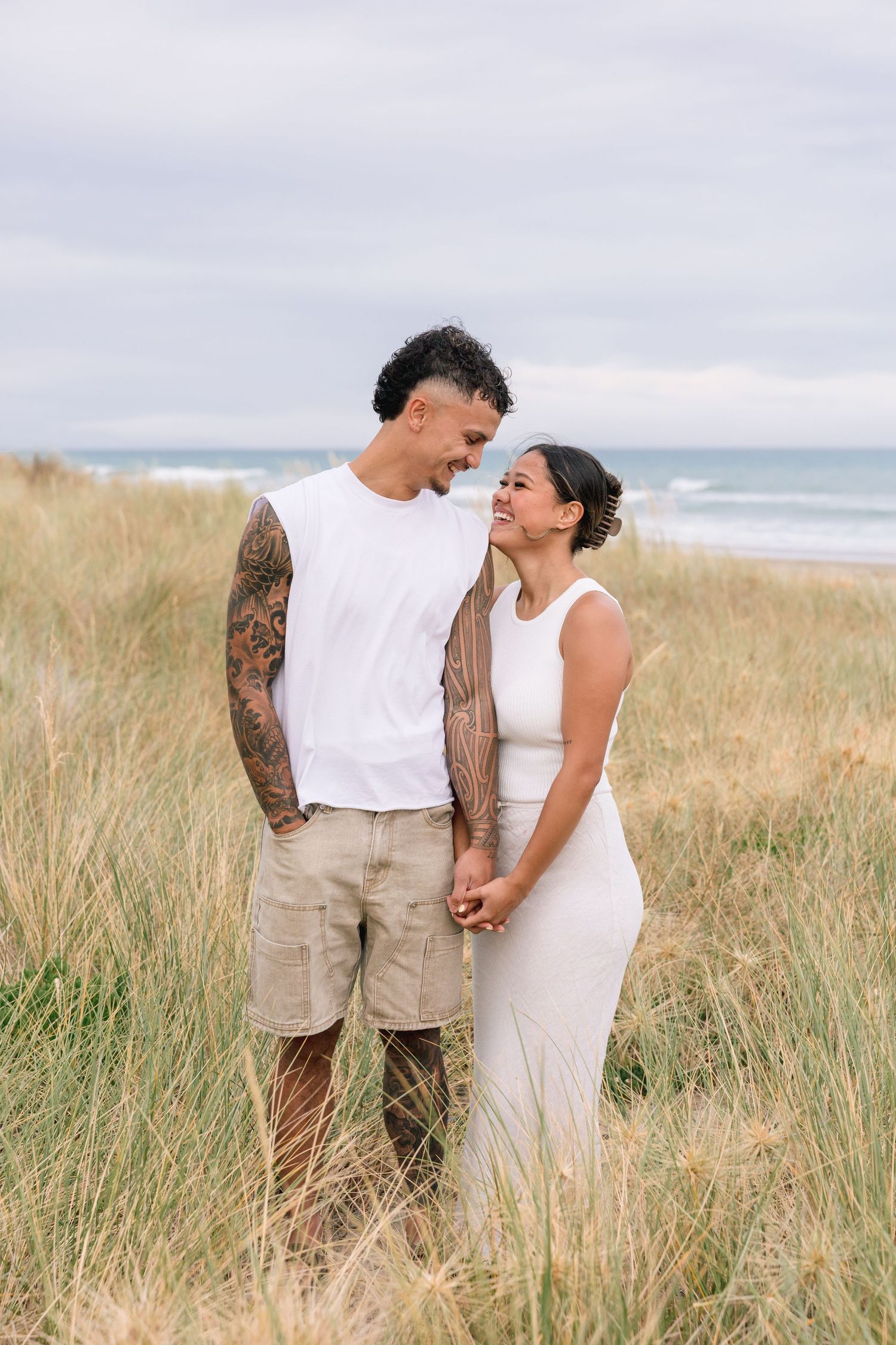Romantic couple embracing in tall grass on a cloudy beach during a photoshoot.