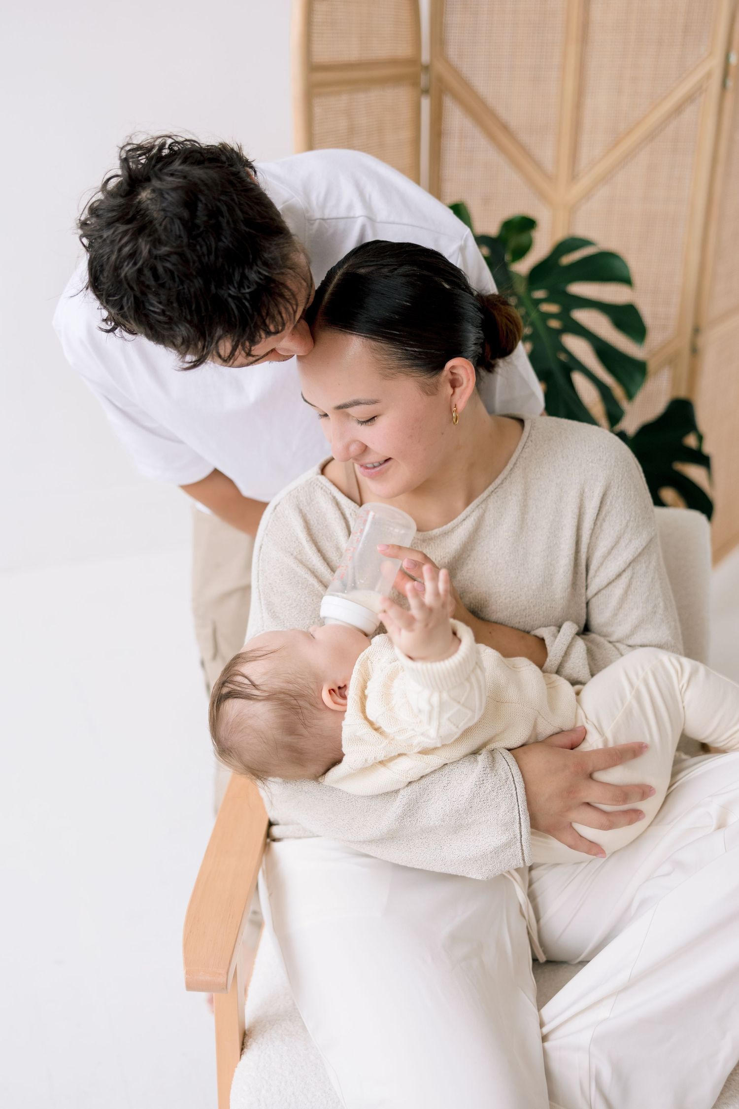 A smiling mother bottle-feeds her baby while a man leans in to watch, seated in a bright, minimalist room.