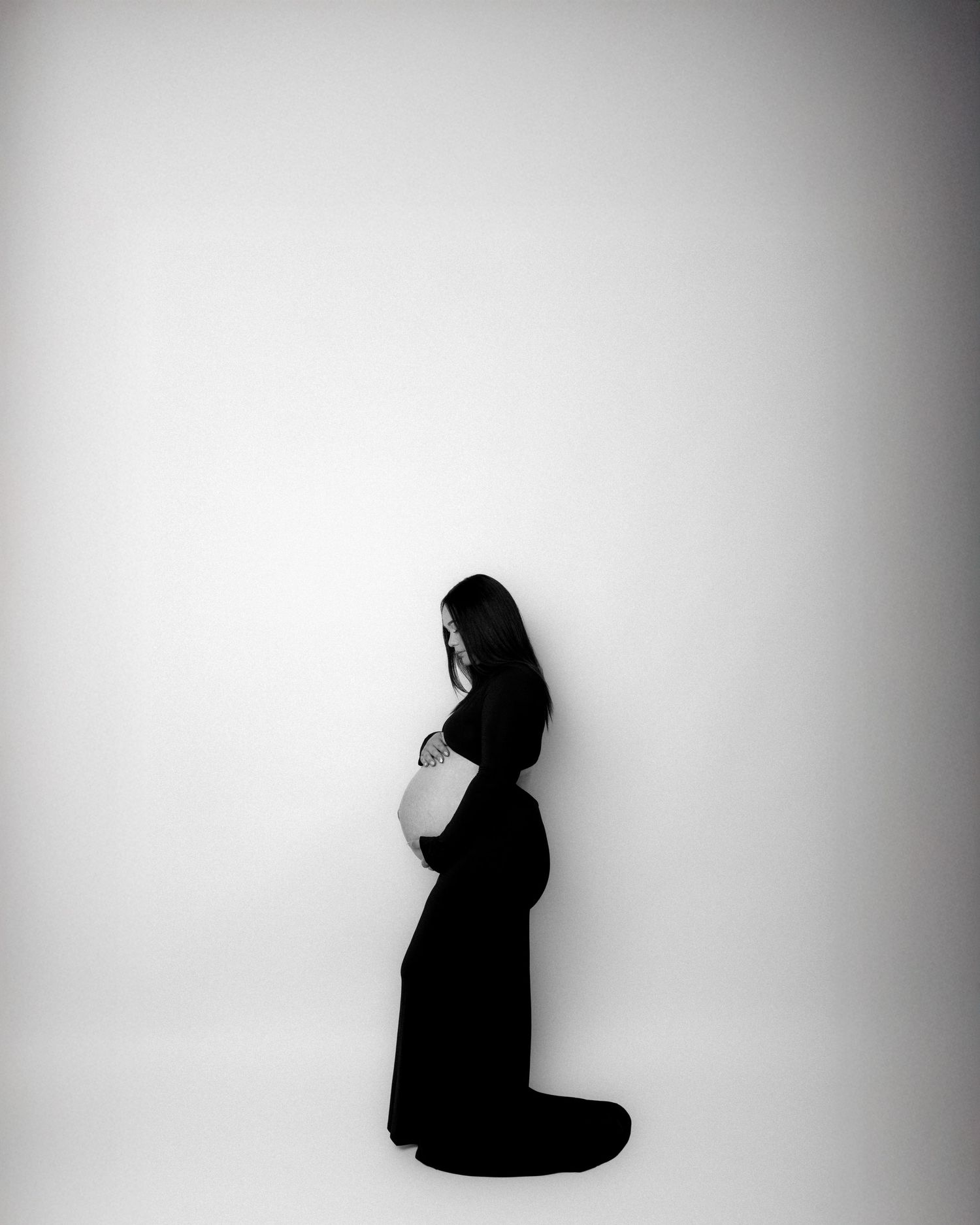 Black and white maternity photo of a pregnant woman in a black gown, cradling her belly in a minimalist studio.