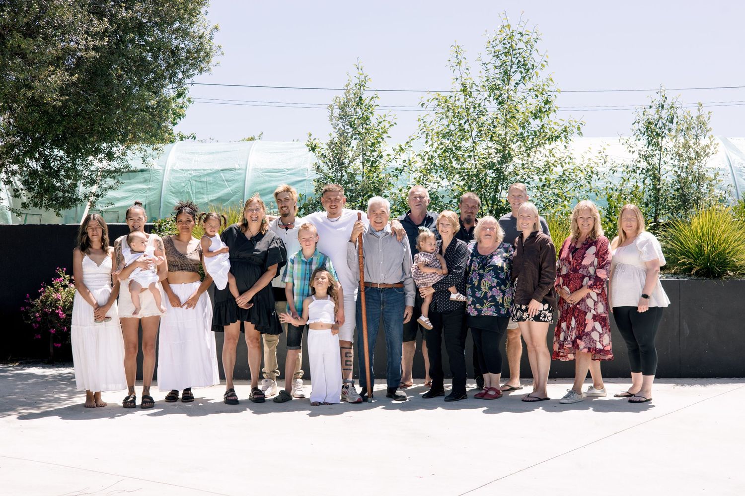 Large family group portrait outdoors on a sunny day, spanning multiple generations, posed together smiling on a patio.