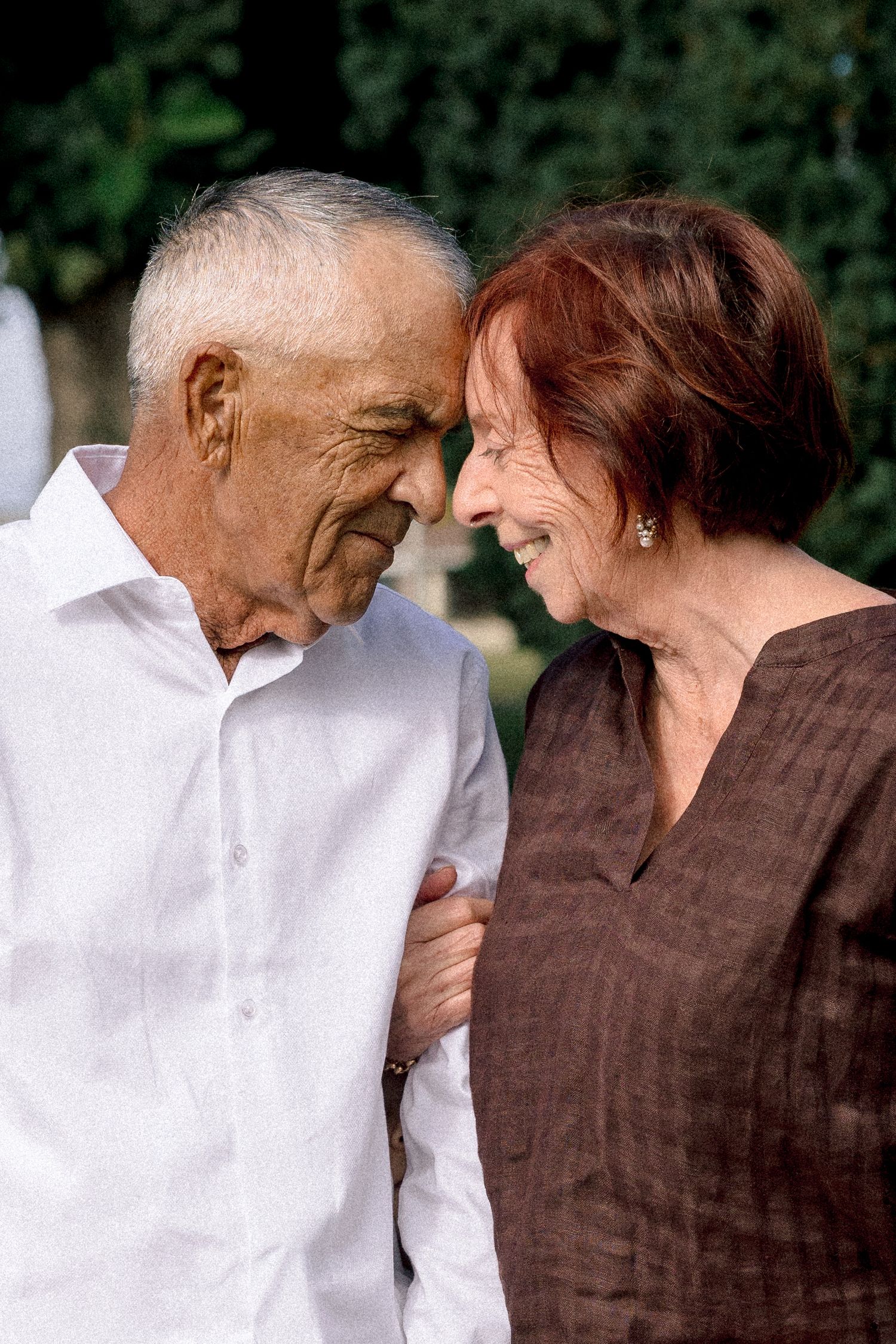 Elderly couple sharing a tender moment outdoors, smiling and touching foreheads in a loving embrace.