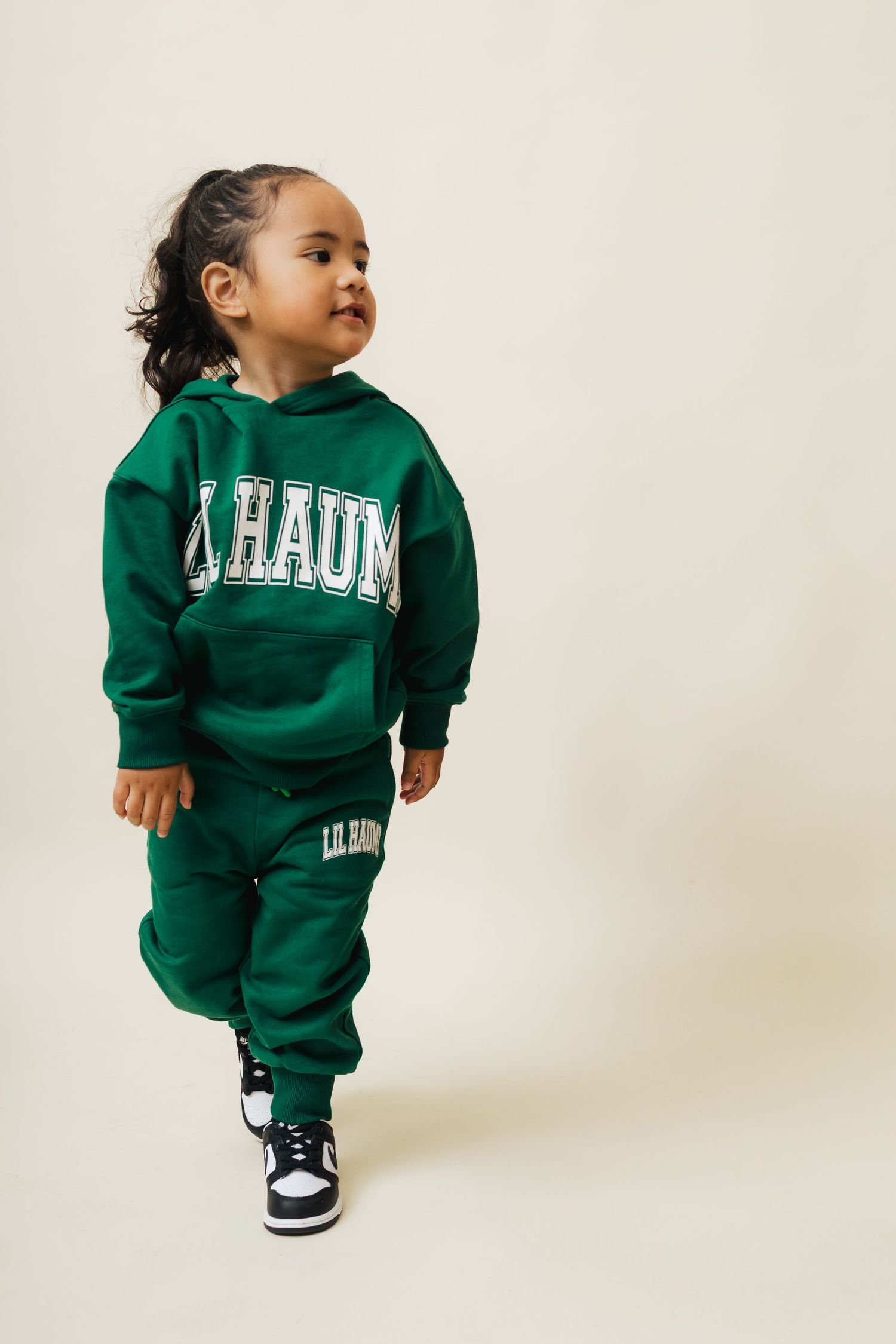 A small child wearing a green university sweatsuit and black sneakers poses against a white background.