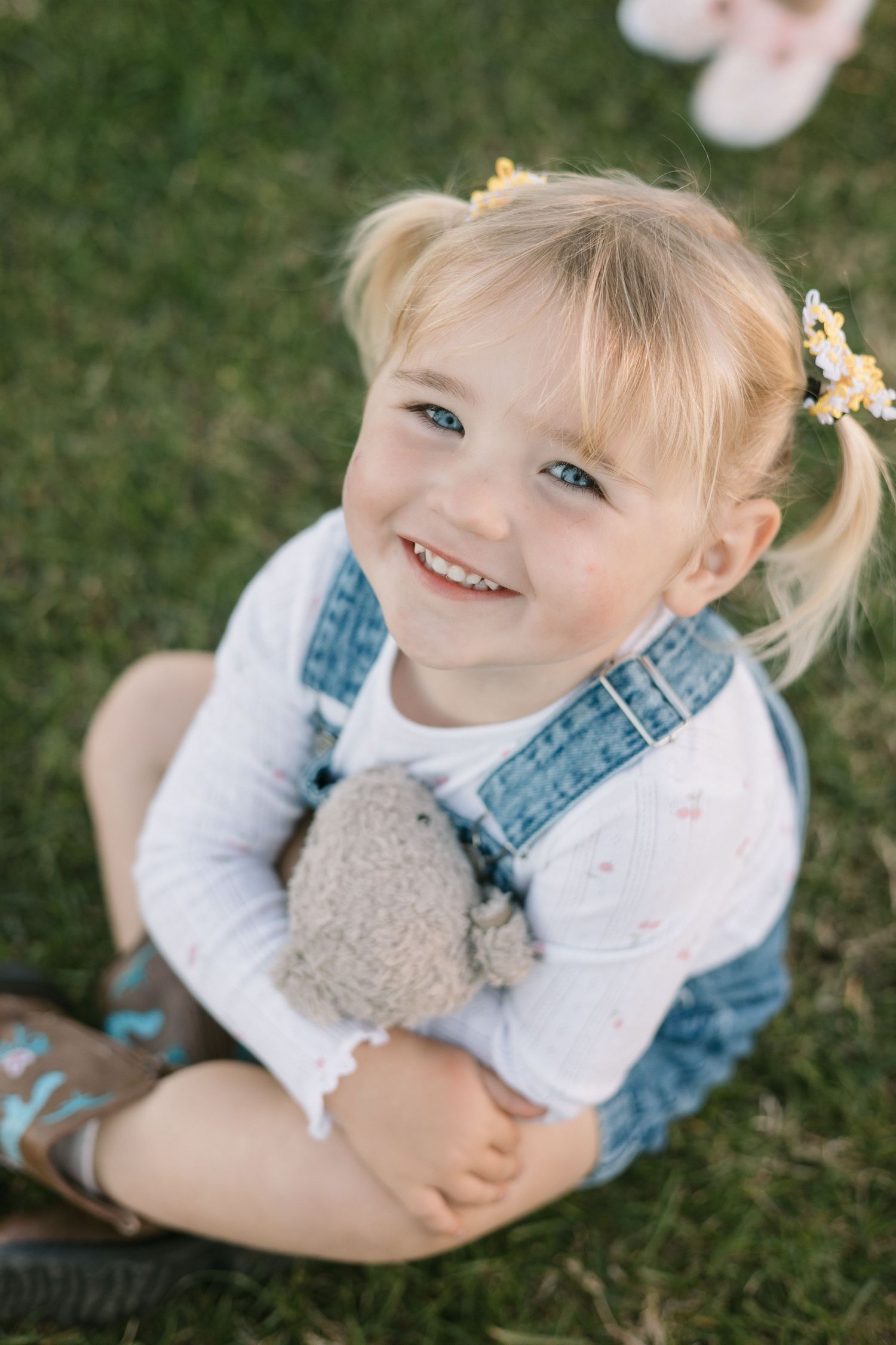 A child in denim overalls and white shirt sits on grass while holding a stuffed toy.