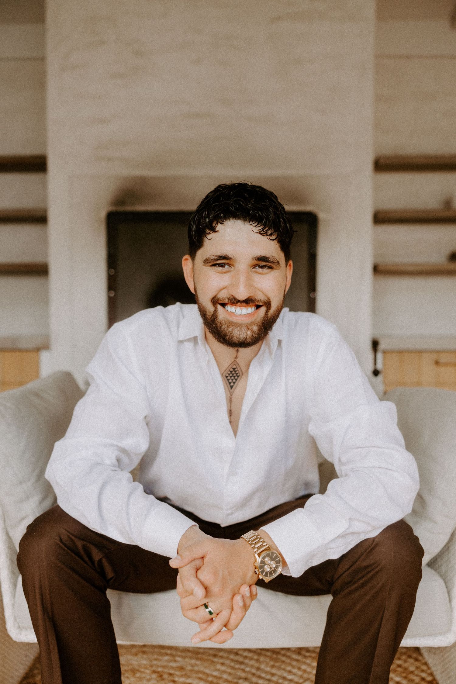Person in white shirt and brown pants smiling while sitting casually in a minimalist white room.