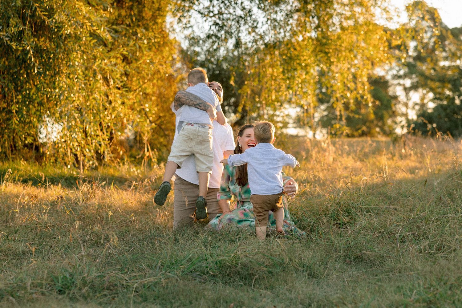 Family enjoying golden hour outdoors, with parents lifting children in a sunny meadow surrounded by trees.