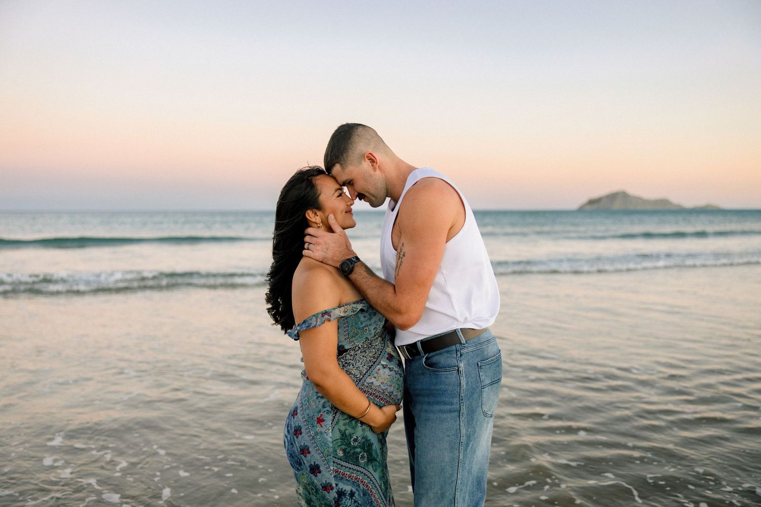 A romantic couple embraces on a beach at sunset with gentle waves and mountains in the background.
