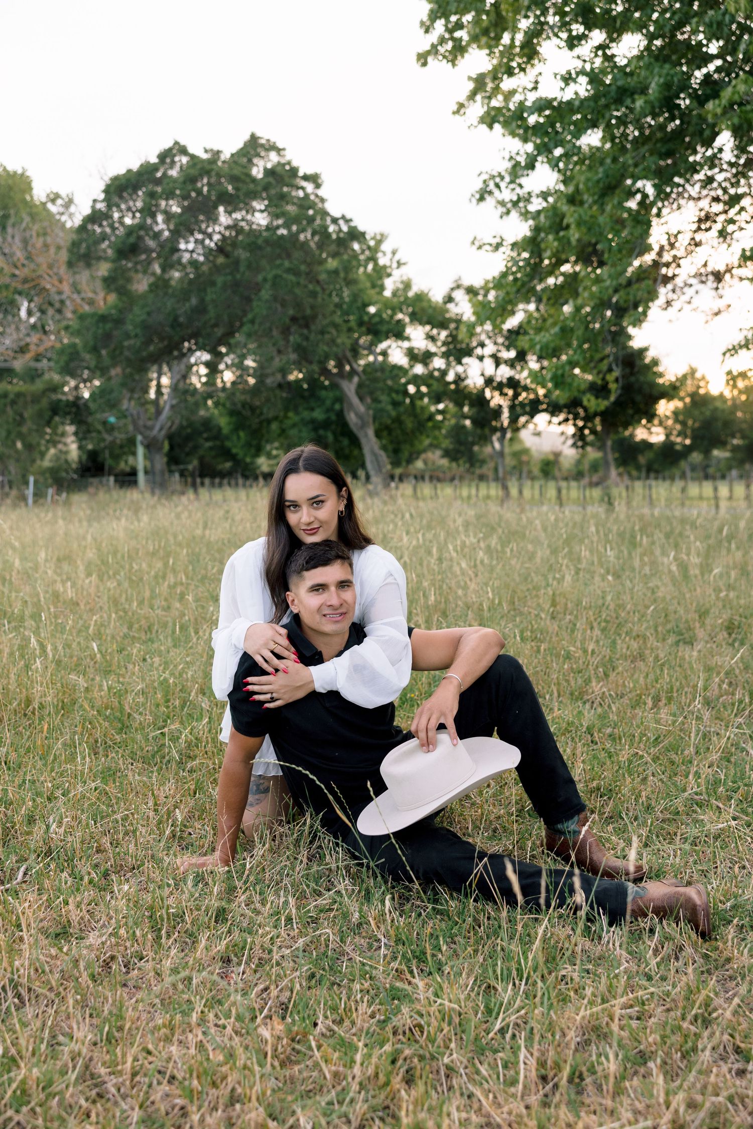 A couple relaxes together on grassy field during golden hour with trees in background.