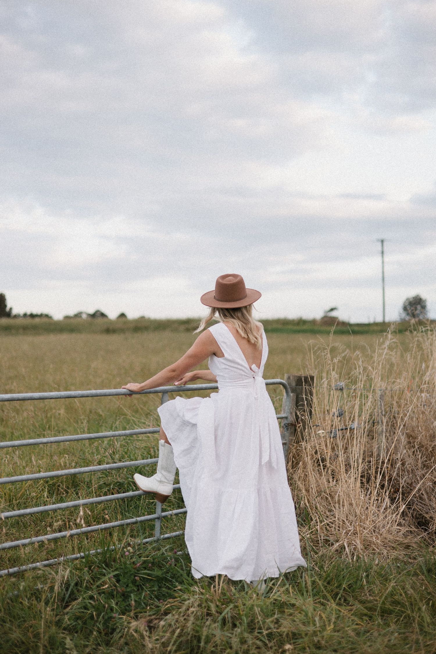 A person in a flowing white dress and brown hat leans against a metal fence in a rural field setting.