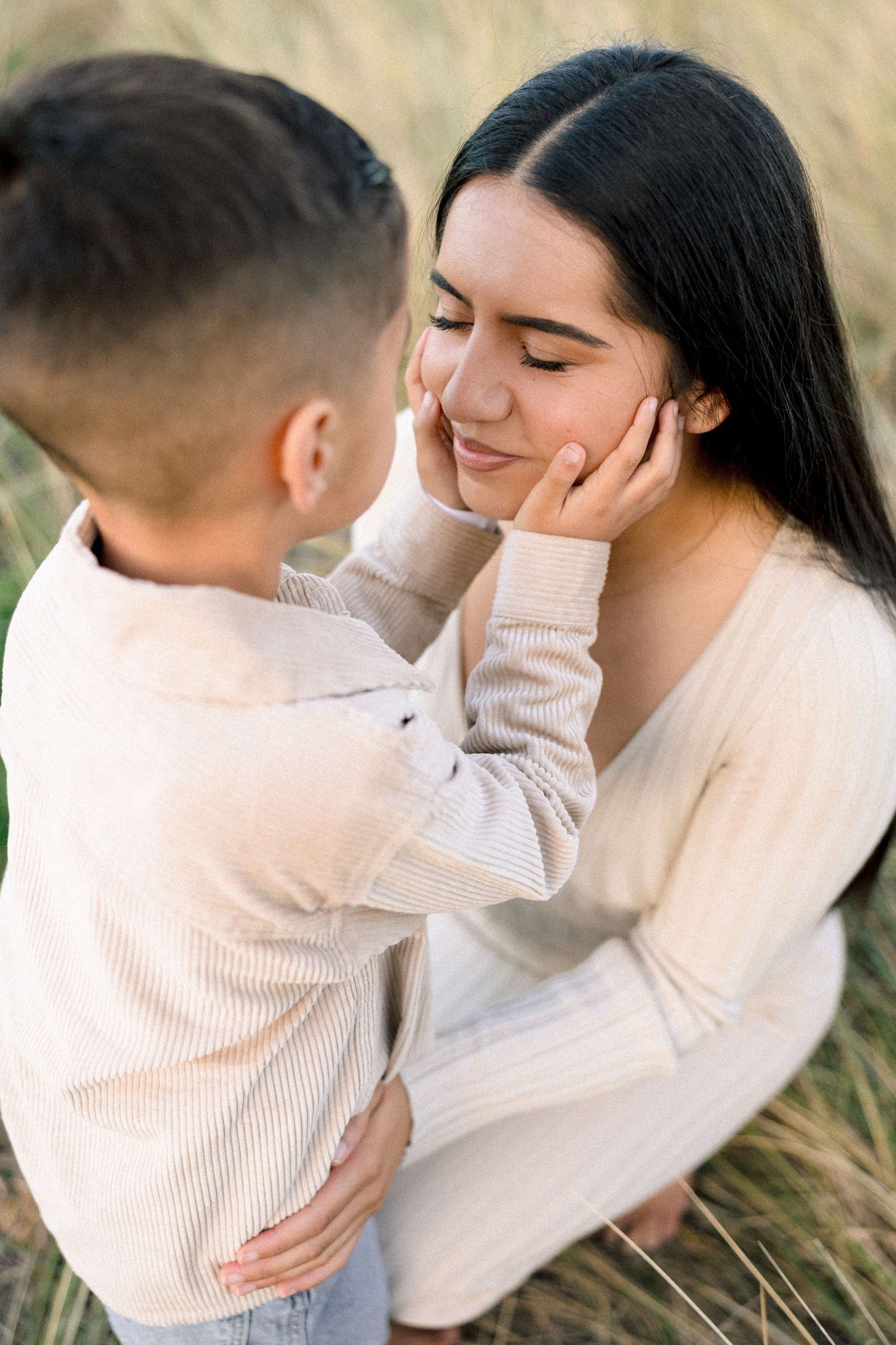 A young boy tenderly cups a smiling woman's face in a sunlit grassy field, sharing a sweet moment.
