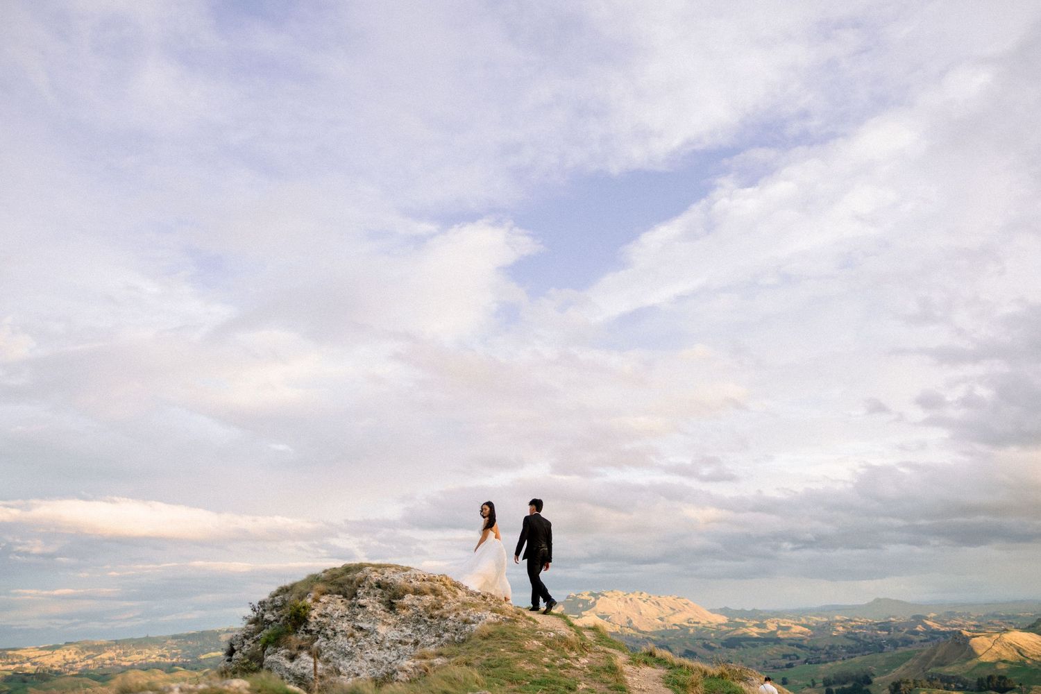 Couple standing on a rocky mountain peak with vast cloudy sky and scenic landscape in the background.