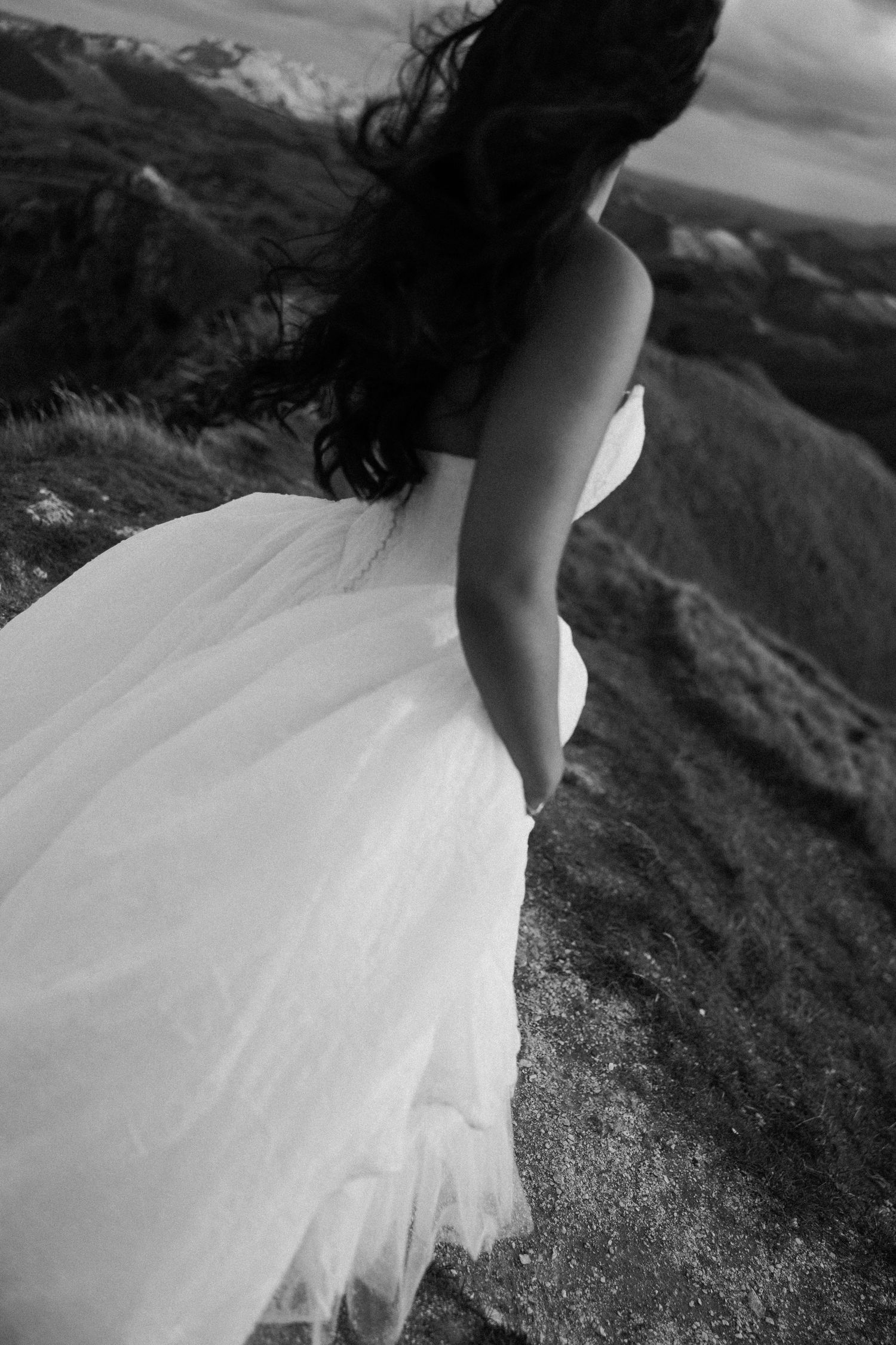 Black and white photo of a bride in a flowing white gown standing on a rocky cliff, her dark hair blowing in the wind.