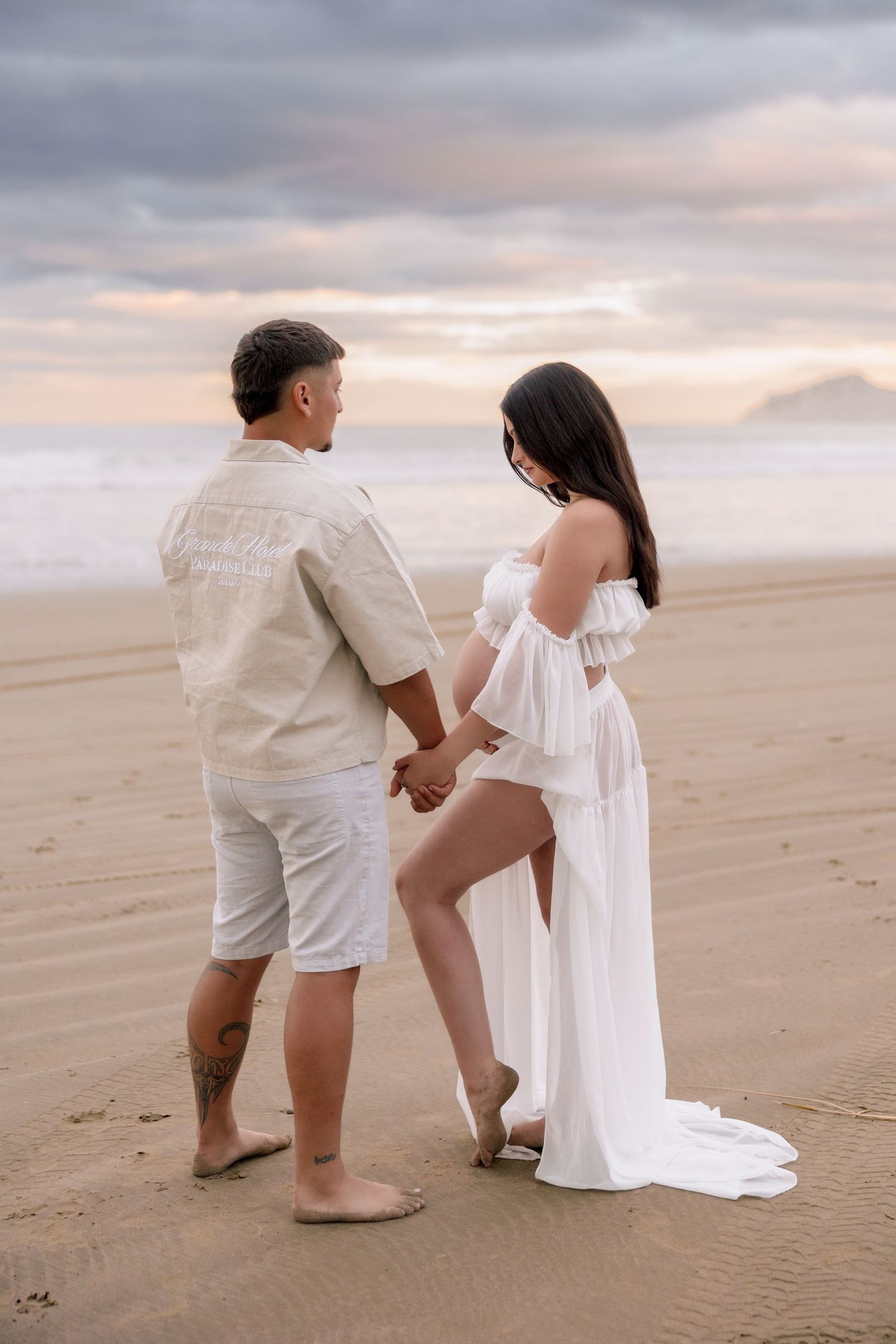 A couple in white outfits walks hand in hand along a sandy beach at sunset creating a romantic silhouette.