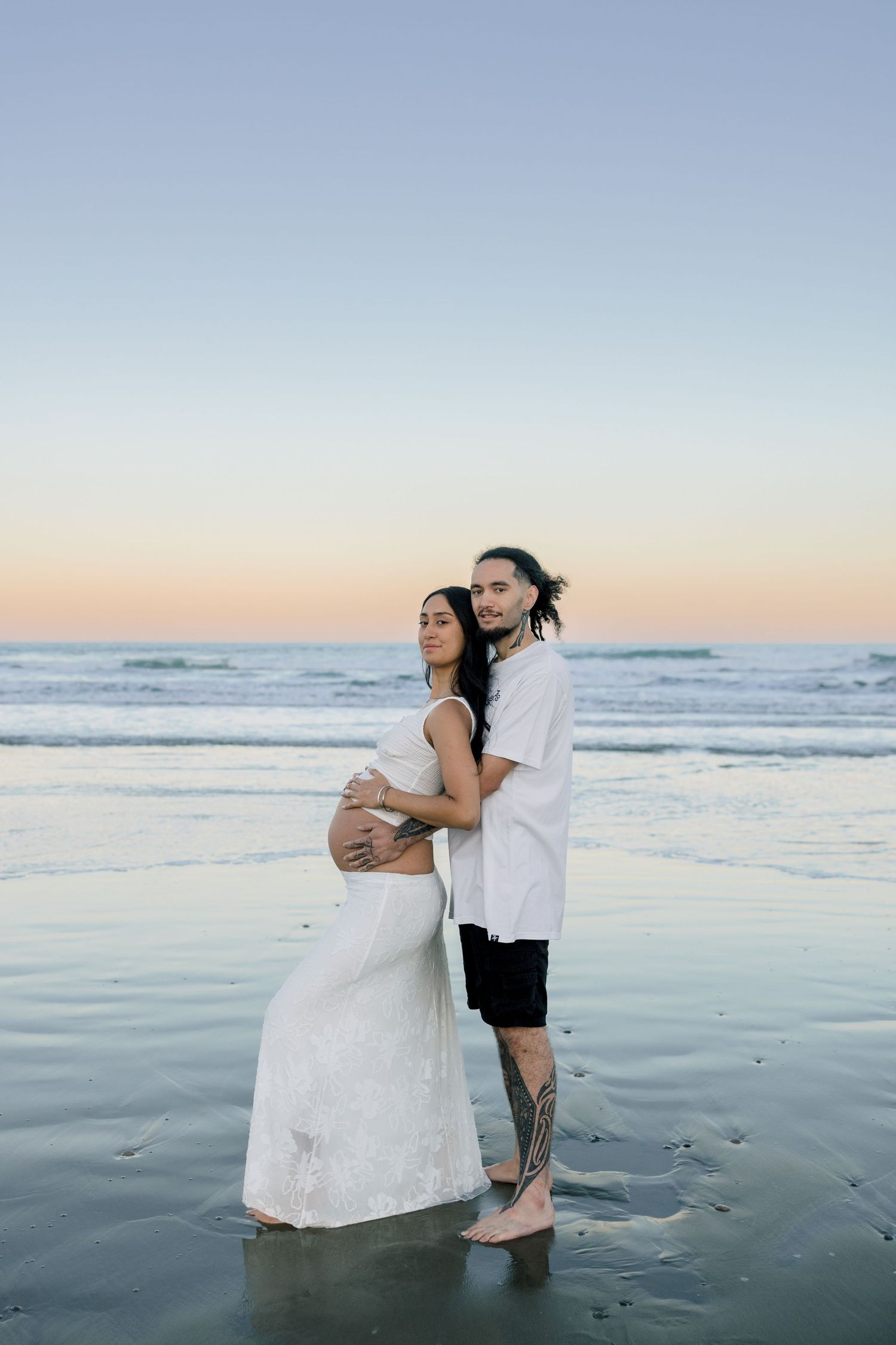 Pregnant couple embracing on beach at sunset, woman in white dress, man with tattoos, ocean waves behind them.