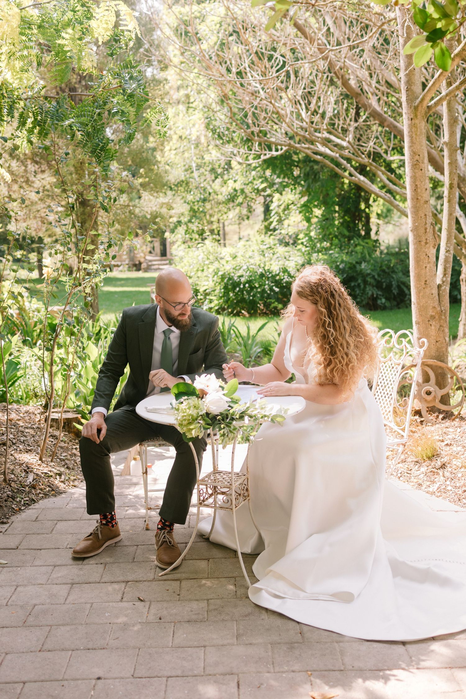 Garden wedding scene with couple seated at white table surrounded by greenery and flowers.