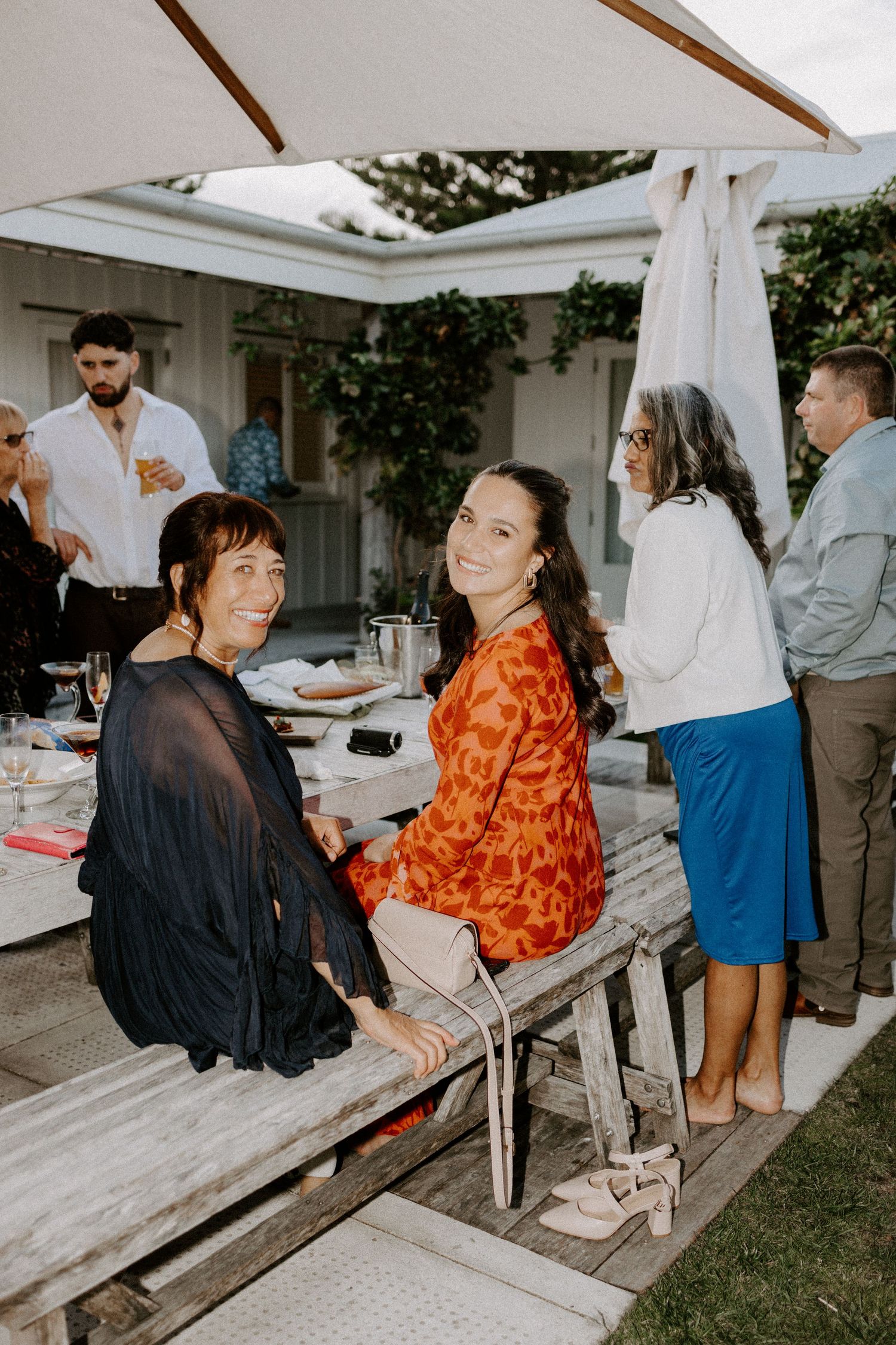 Wedding guests socializing at an outdoor reception with white tent and wooden benches.
