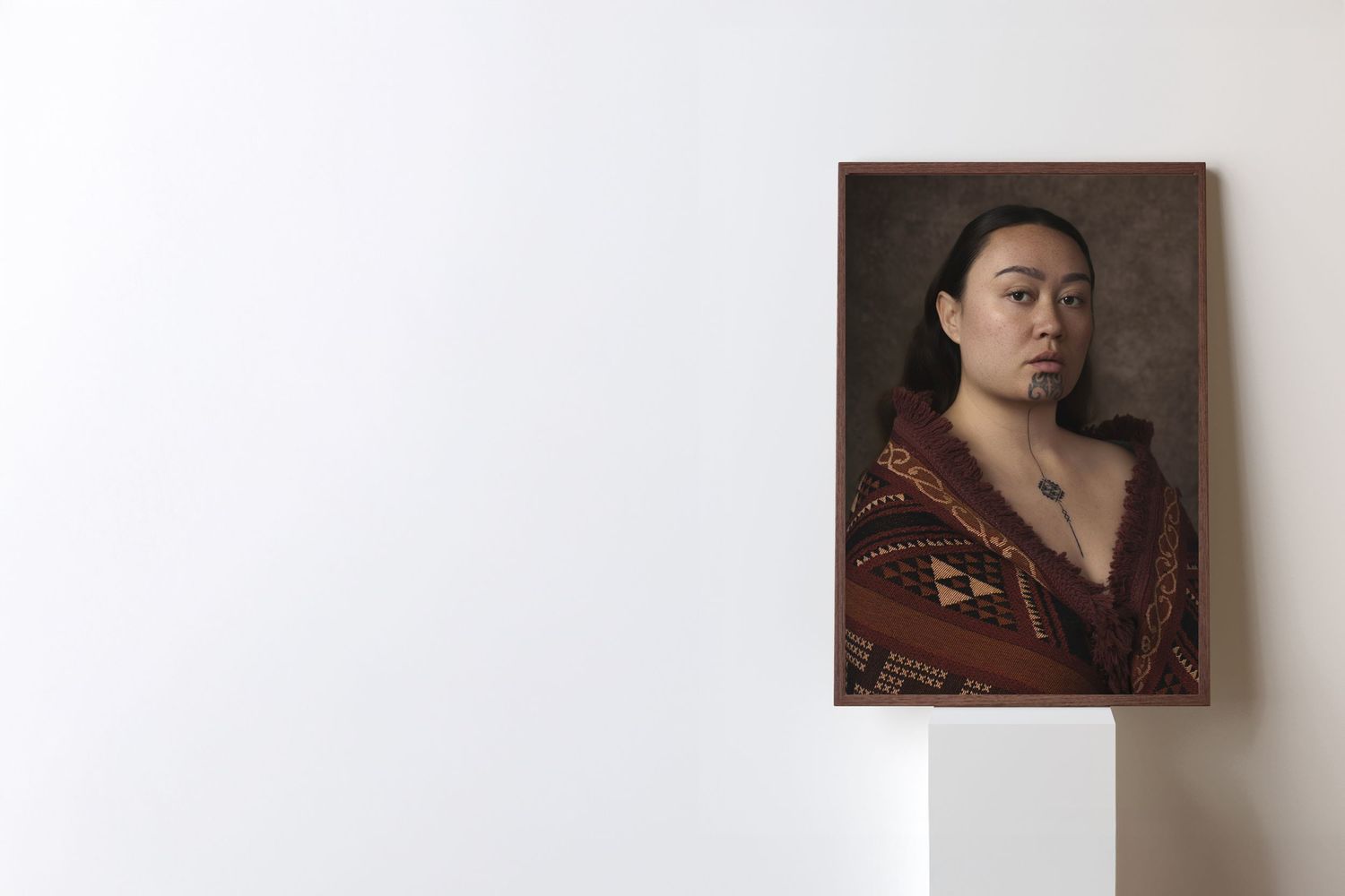 Framed portrait painting of a young Asian woman in a red patterned shawl, displayed on a white pedestal against a white wall.