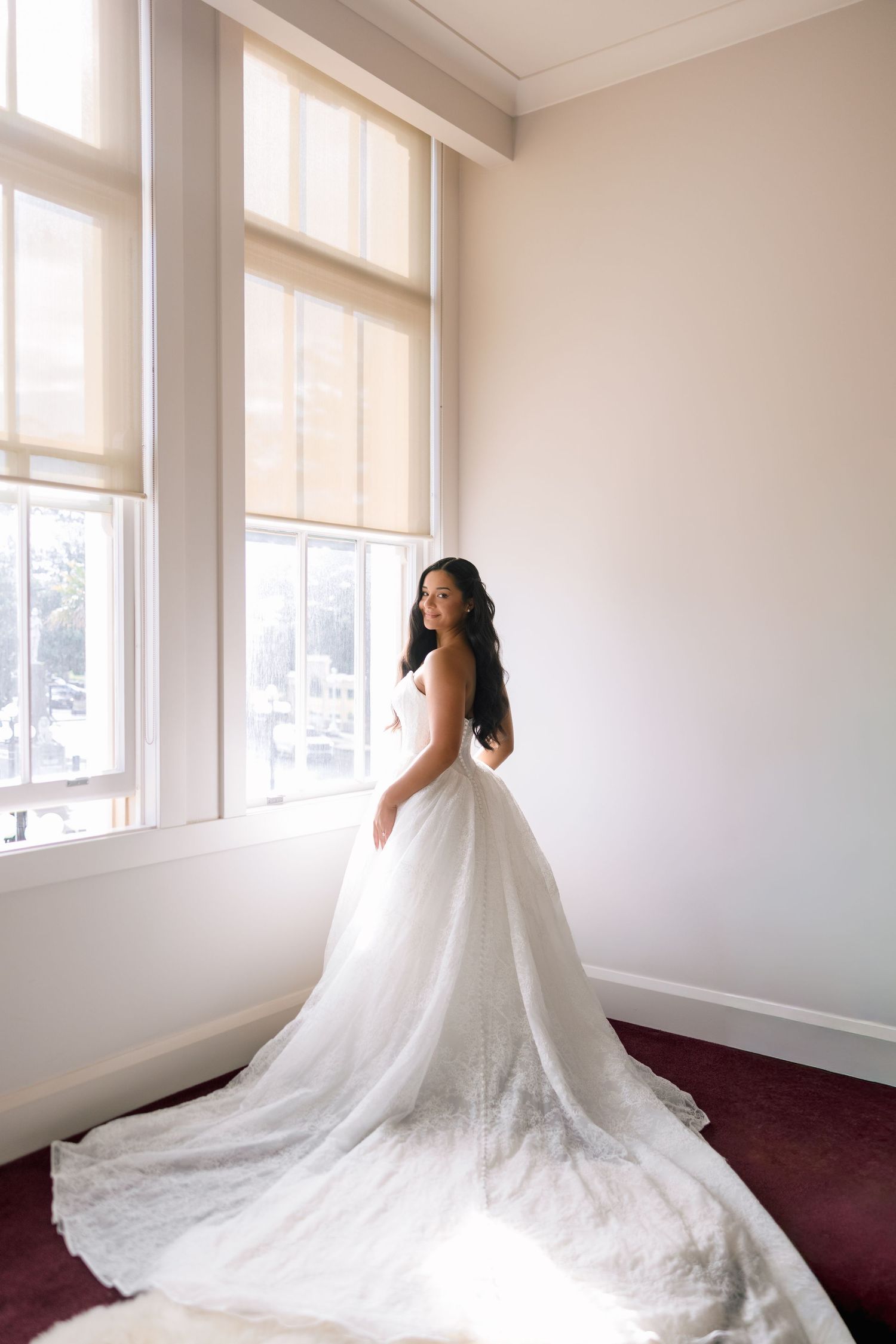 Bride in a strapless white ballgown with a long train stands by large windows in a sunlit room.