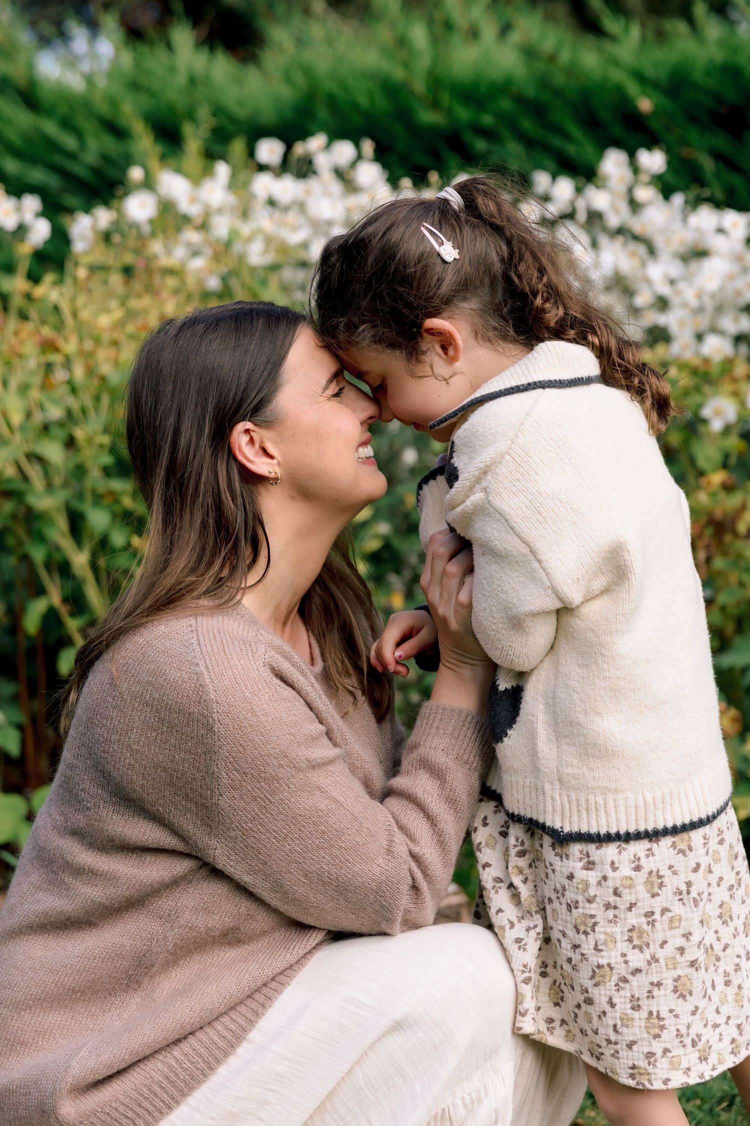 A mother and young daughter share a tender nose-to-nose moment in a lush garden with white flowers.