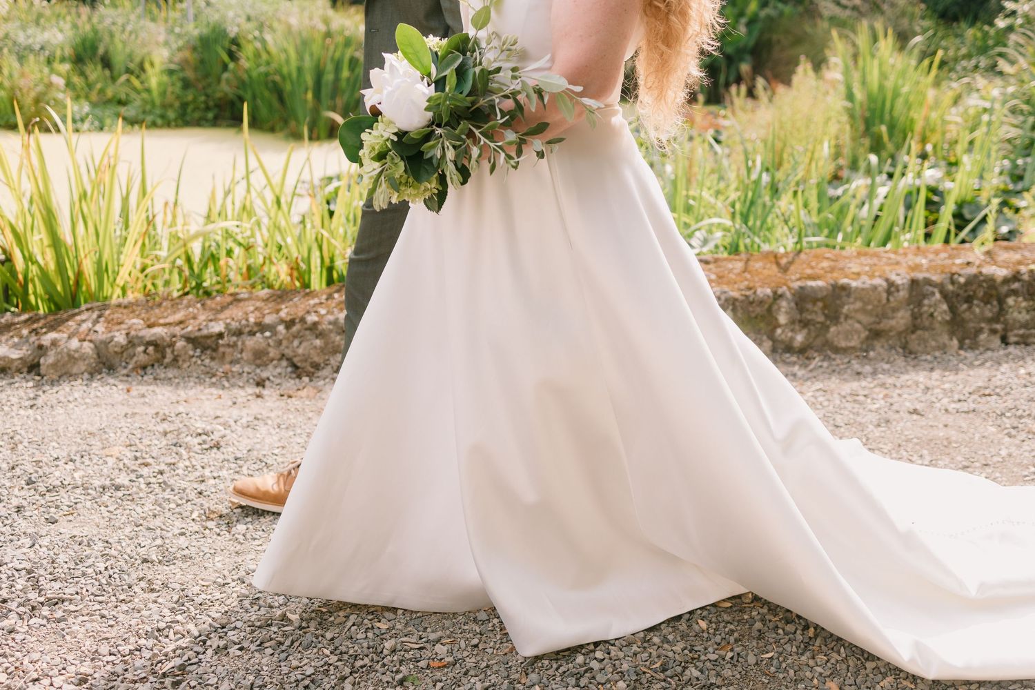 White wedding dress train sweeping across stone pathway in garden setting.