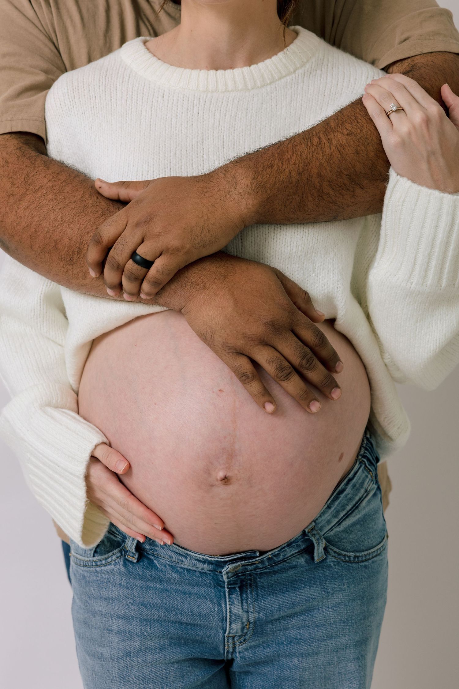 A pregnant woman in a white sweater and jeans with her partner's hands cradling her bare baby bump.