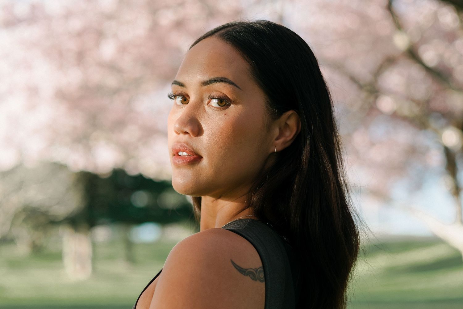 A woman with dark hair poses outdoors against a backdrop of blooming cherry blossom trees in springtime.