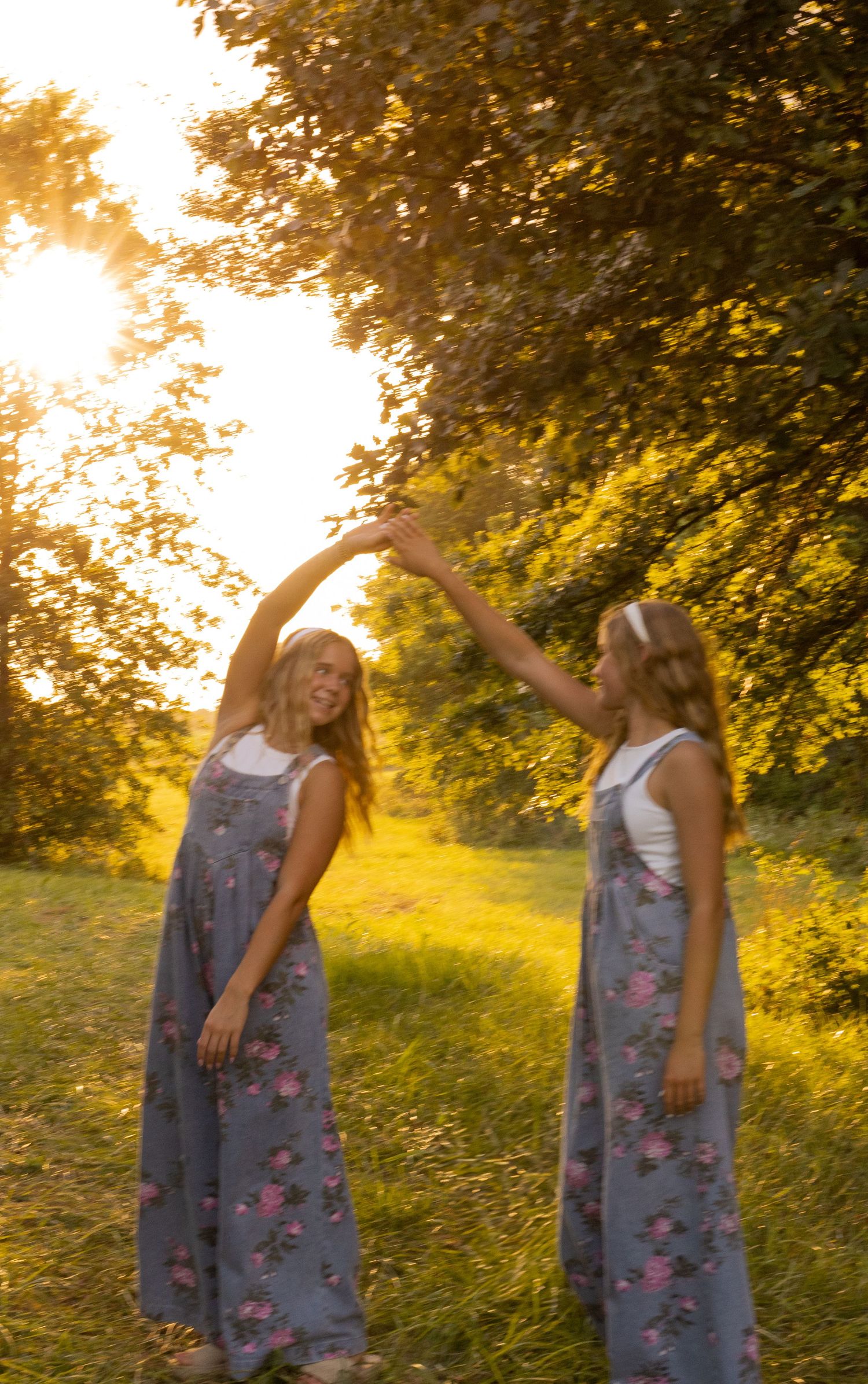 Two girls in matching floral overalls dancing under sunlit trees in a lush green field at sunset.