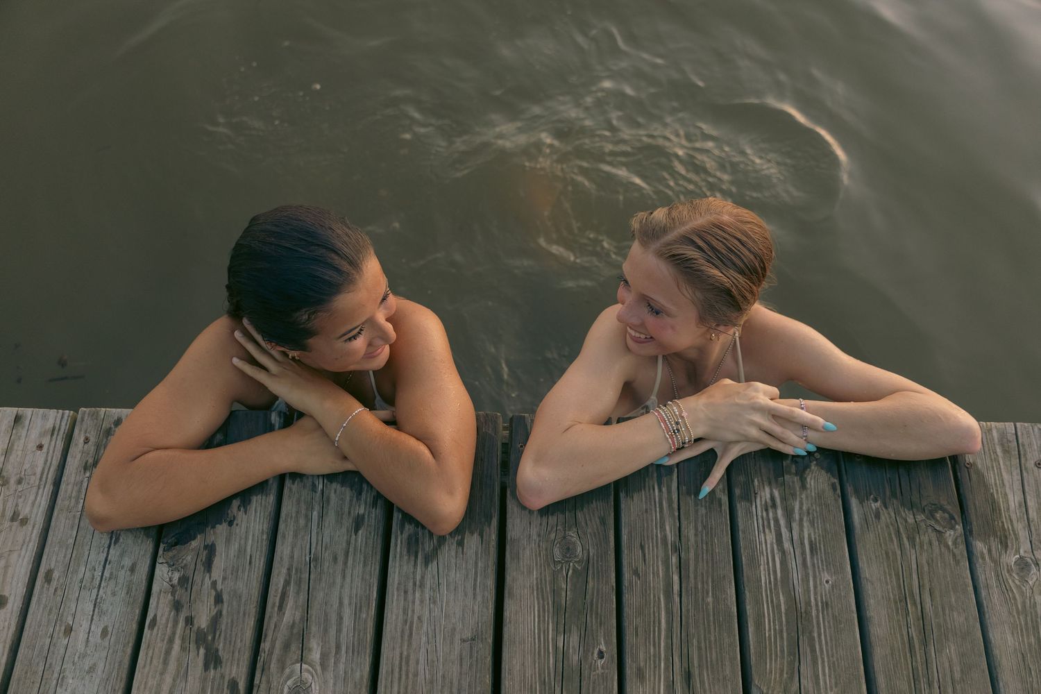 Two women swimming in a lake, resting on a wooden dock, smiling and talking together outdoors.